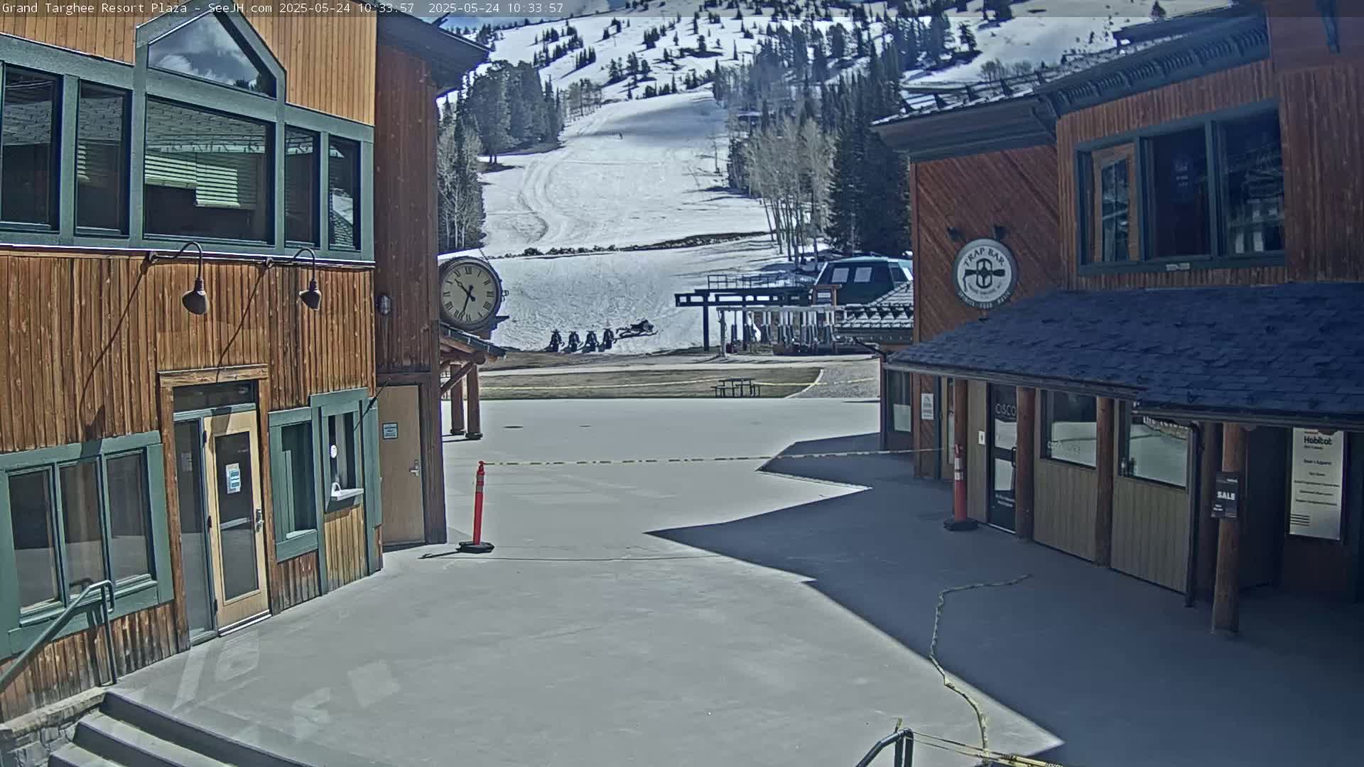 A sunny plaza area between two wooden buildings overlooks a snow-covered ski slope with several snowmobiles parked nearby.