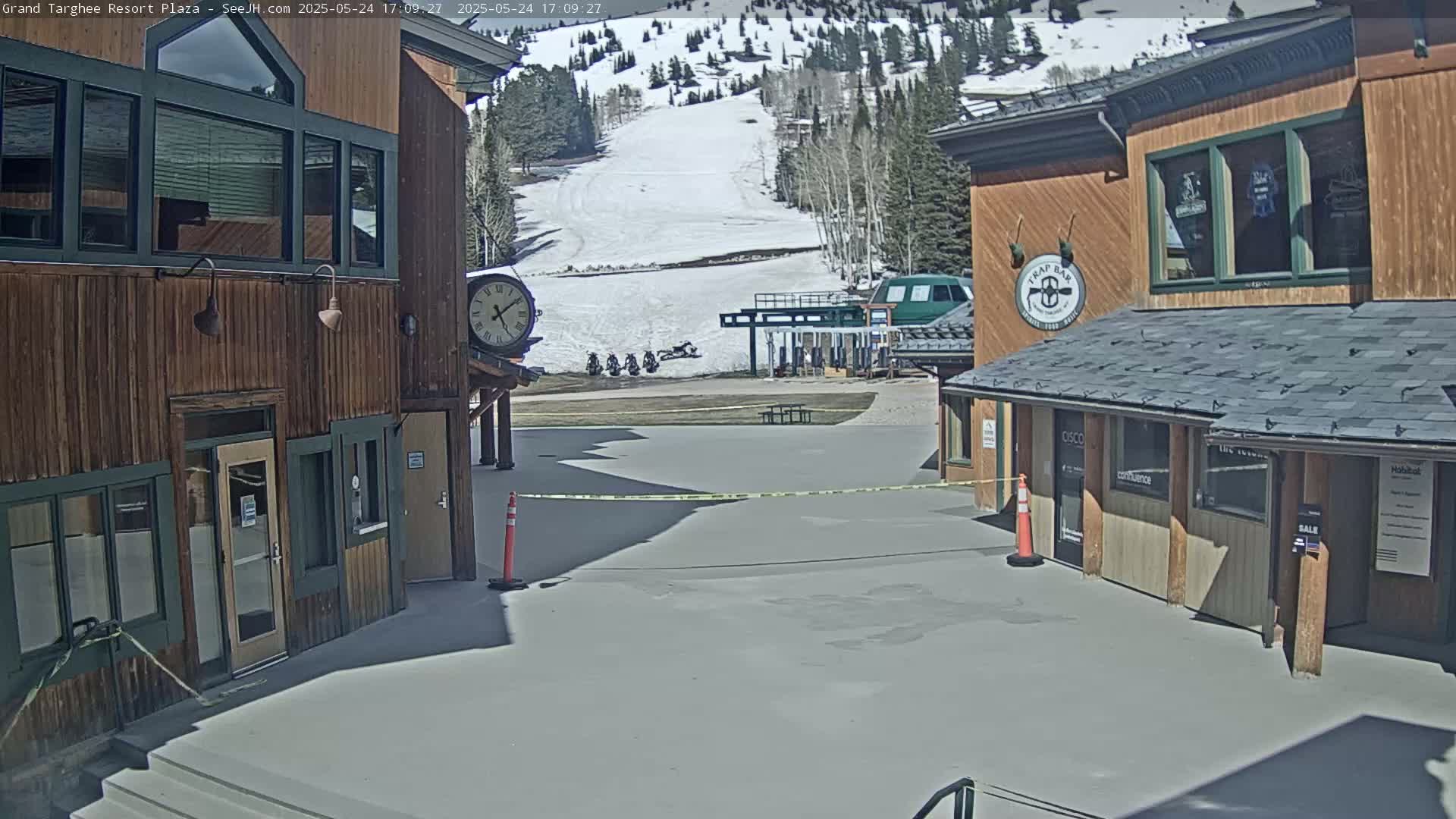 A plaza with two wooden buildings sits before a snow-covered ski slope on a sunny day.