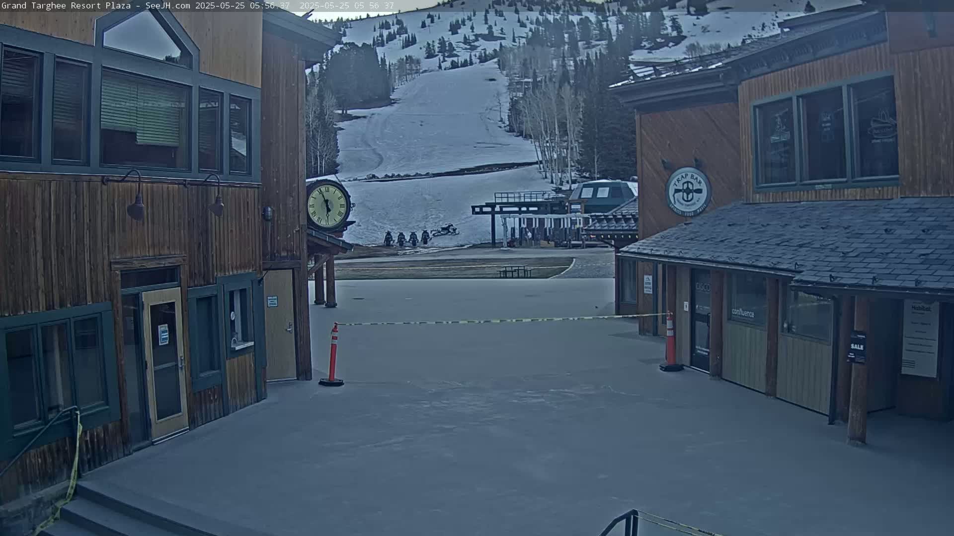 A plaza with two wooden buildings, a clock, and snow-covered slopes in the background under an overcast sky.