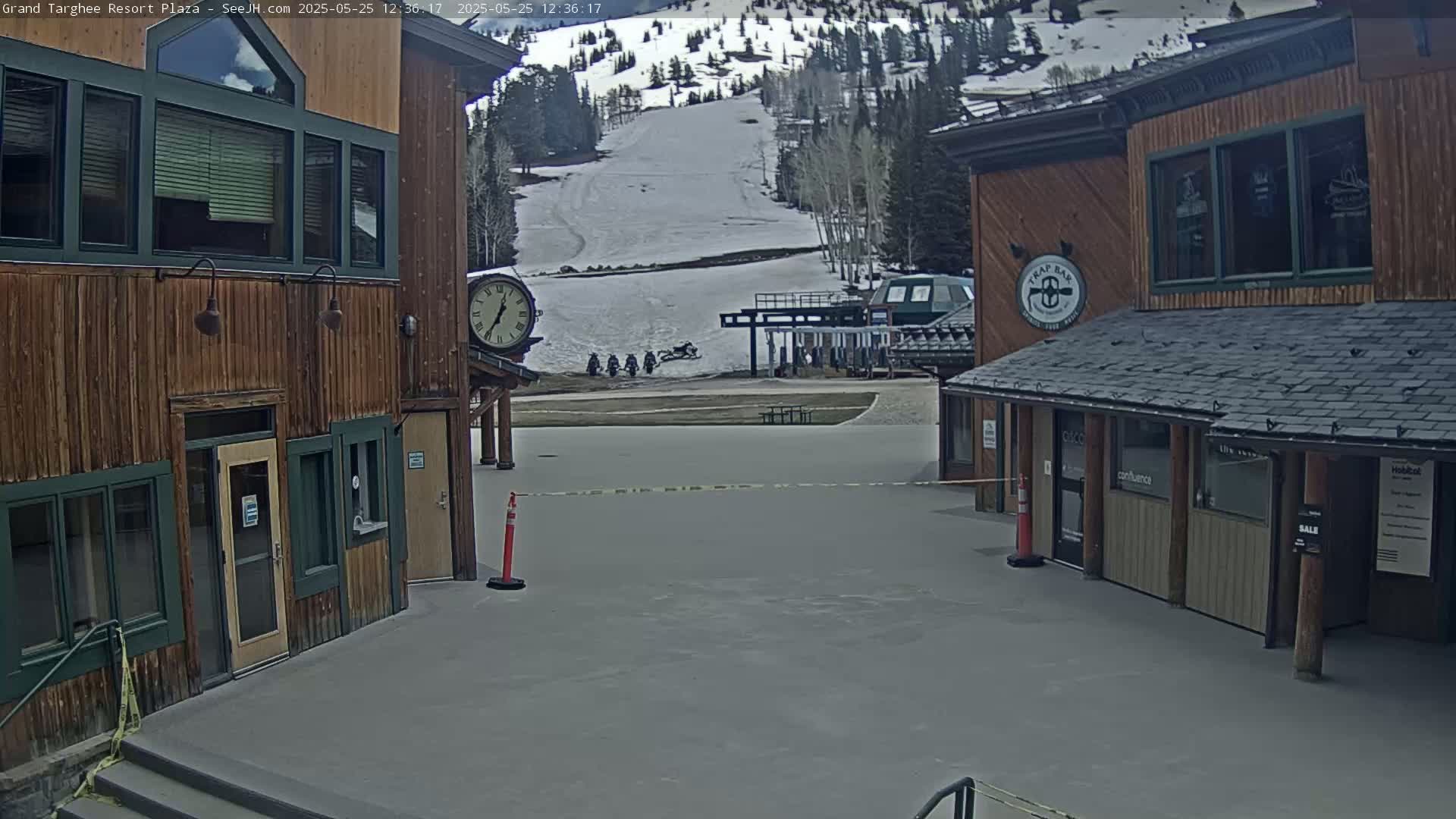 A plaza with two wood-sided buildings overlooks a snow-covered ski slope with a chairlift and several snowmobiles parked below.