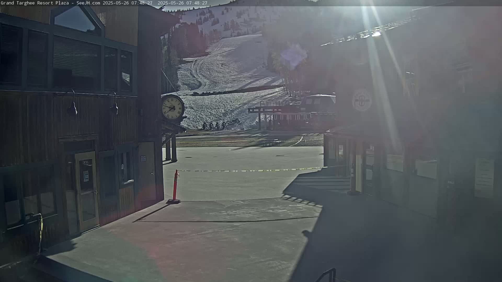 A sunny view from between two buildings shows a snow-covered ski slope in the distance and a mostly empty plaza in the foreground.
