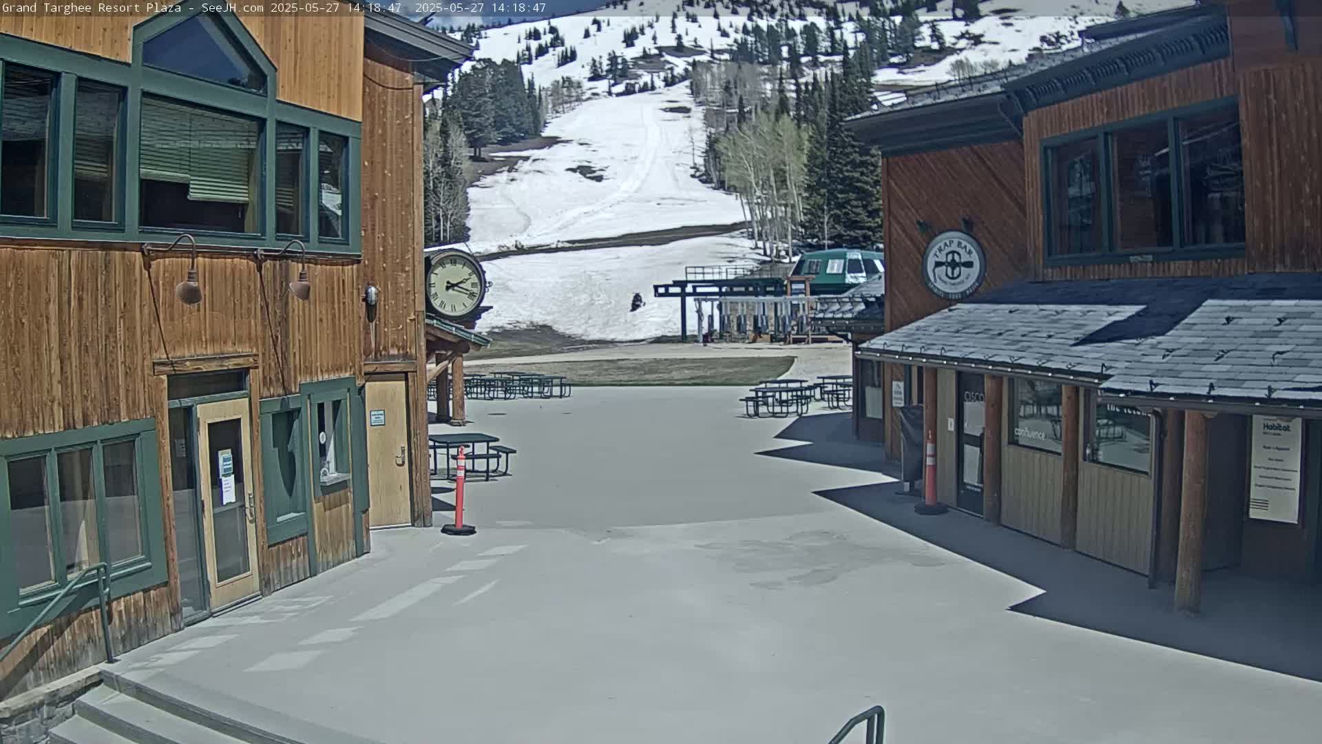 A plaza with two wooden buildings and several picnic tables overlooks a snow-covered ski slope on a sunny day.