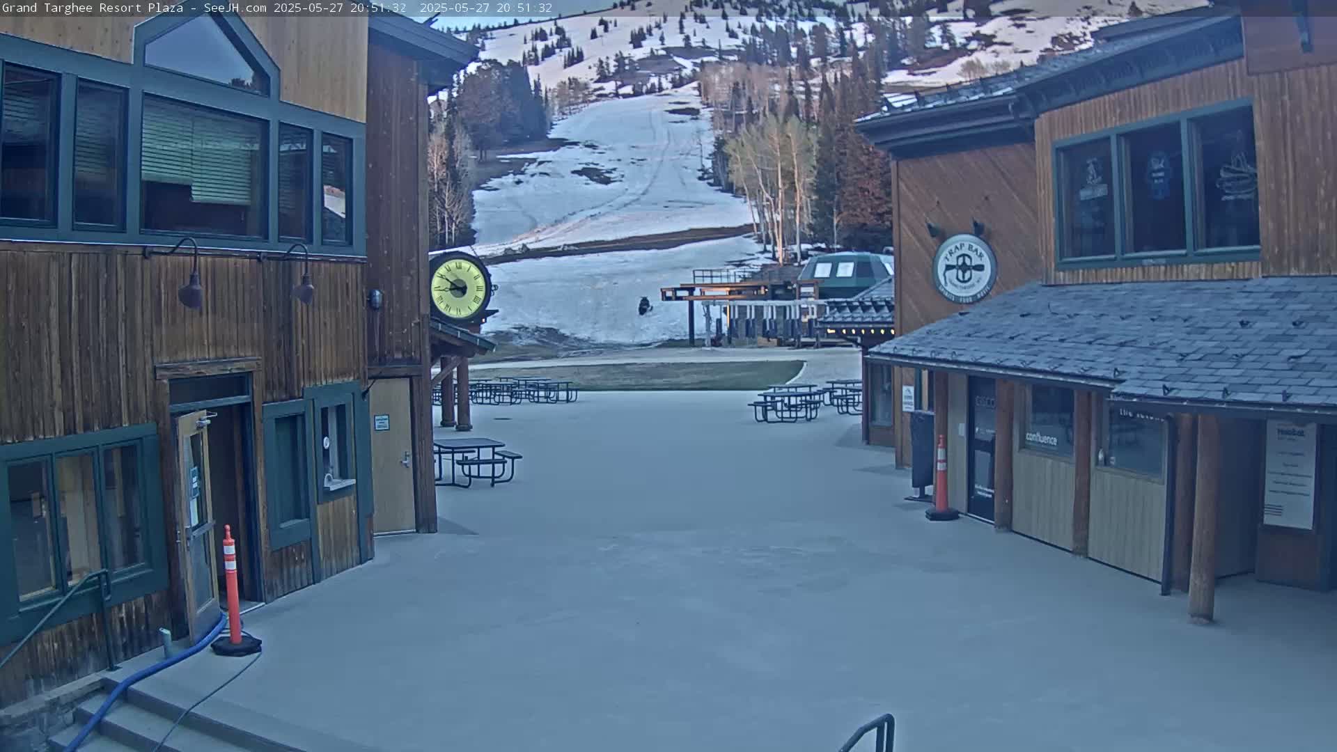 A plaza with wooden buildings and picnic tables sits before a snow-covered ski slope under a twilight sky.