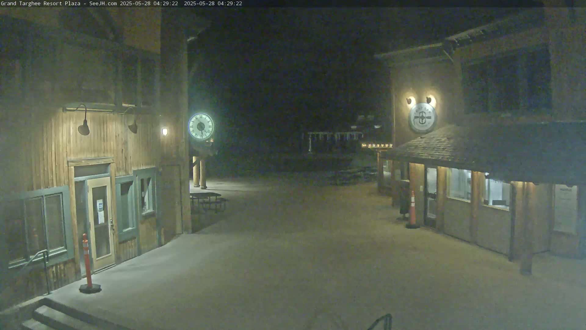 A snow-covered plaza at night with two wooden buildings, one featuring a clock, is illuminated by artificial light.