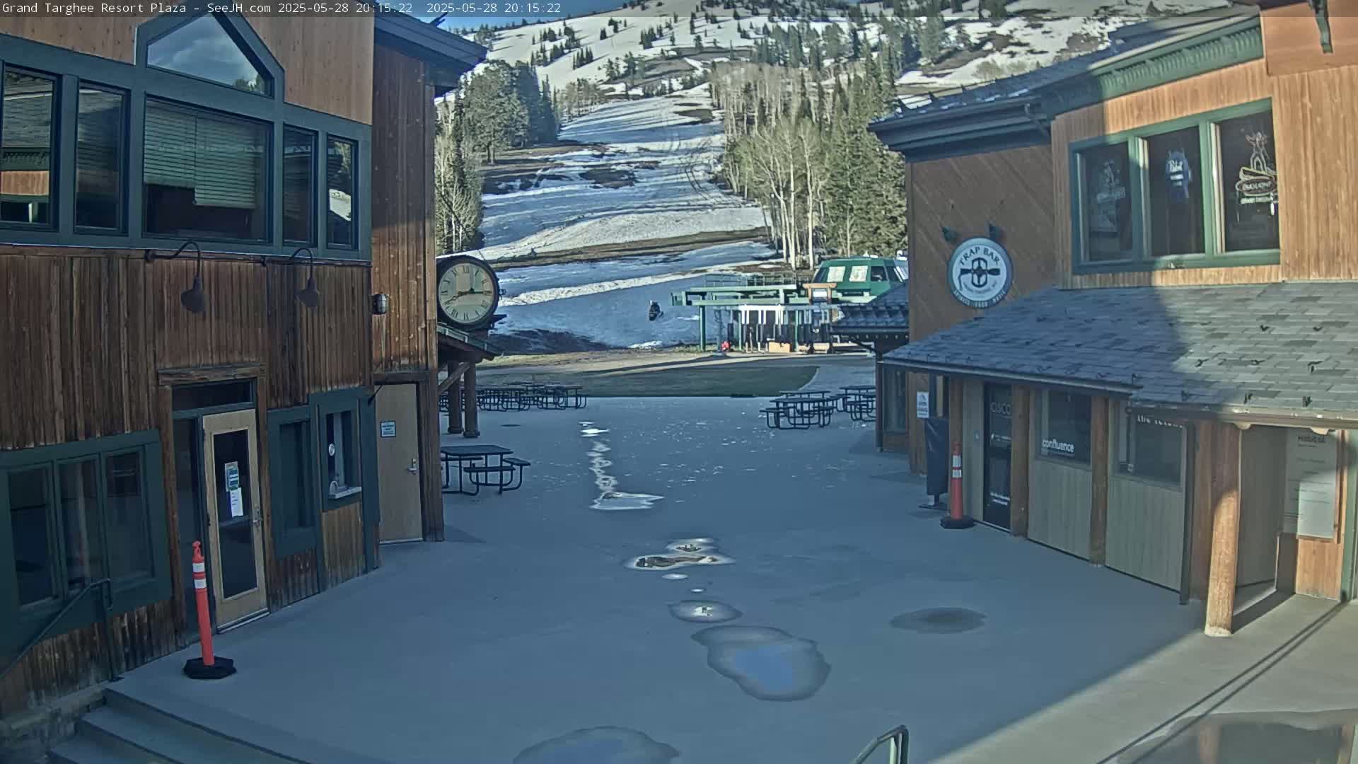 A plaza with wooden buildings, picnic tables, and puddles sits before a snow-covered mountain slope with a ski lift.