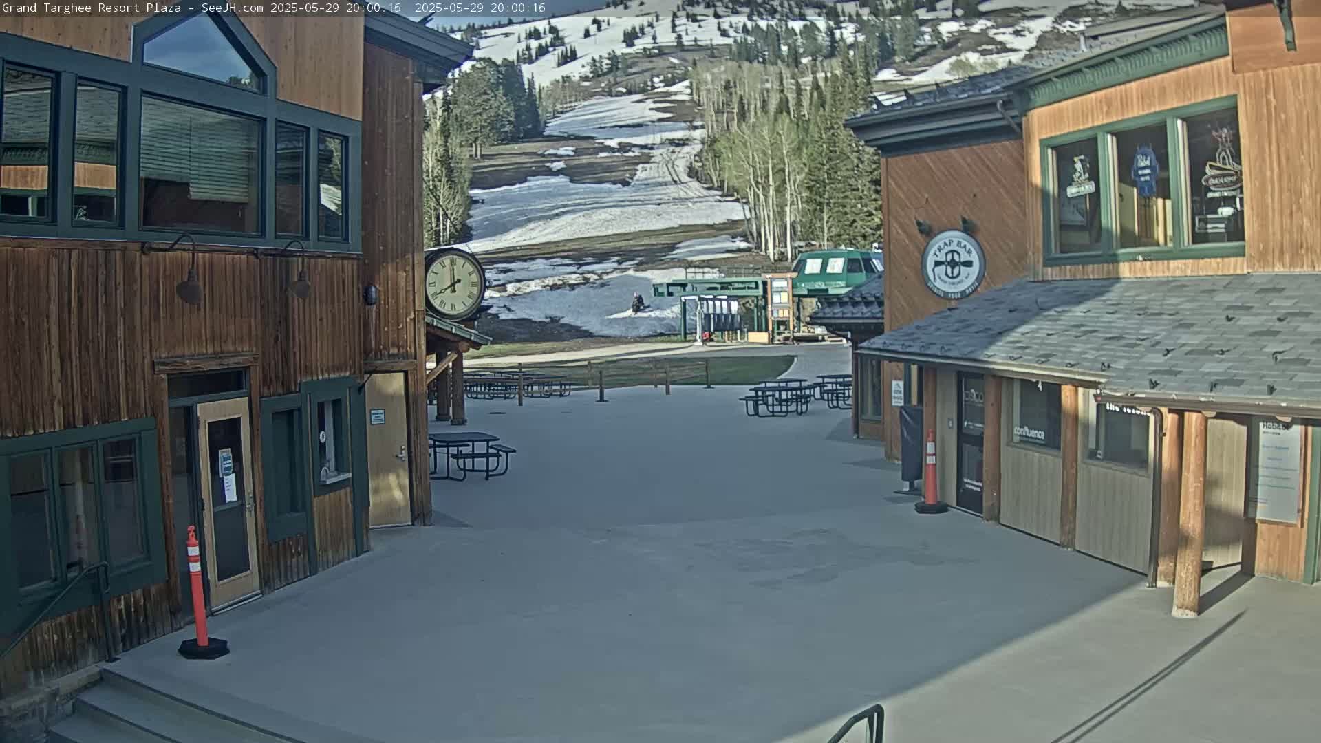 A sunny plaza with several picnic tables sits between two wooden buildings, overlooking a ski slope with patches of snow.