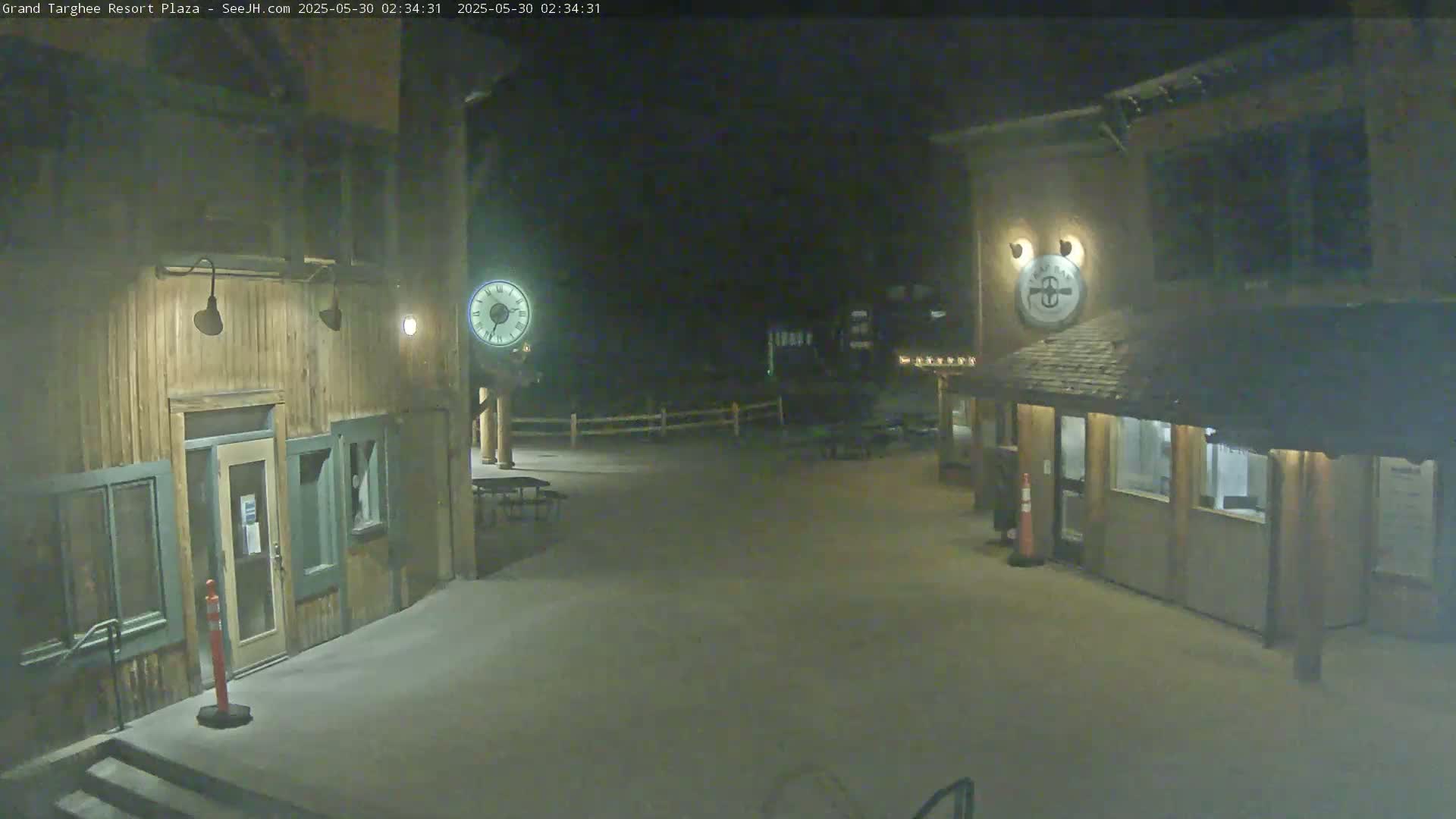 A nighttime view of a plaza with two wooden buildings, outdoor furniture, and clocks on posts.