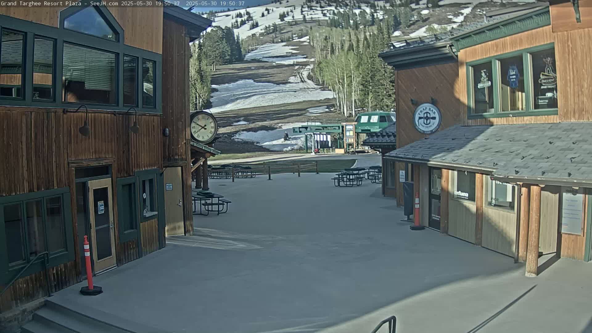 A sunny outdoor plaza with several wooden buildings, picnic tables, and a ski lift visible in the background, which still shows some snow.