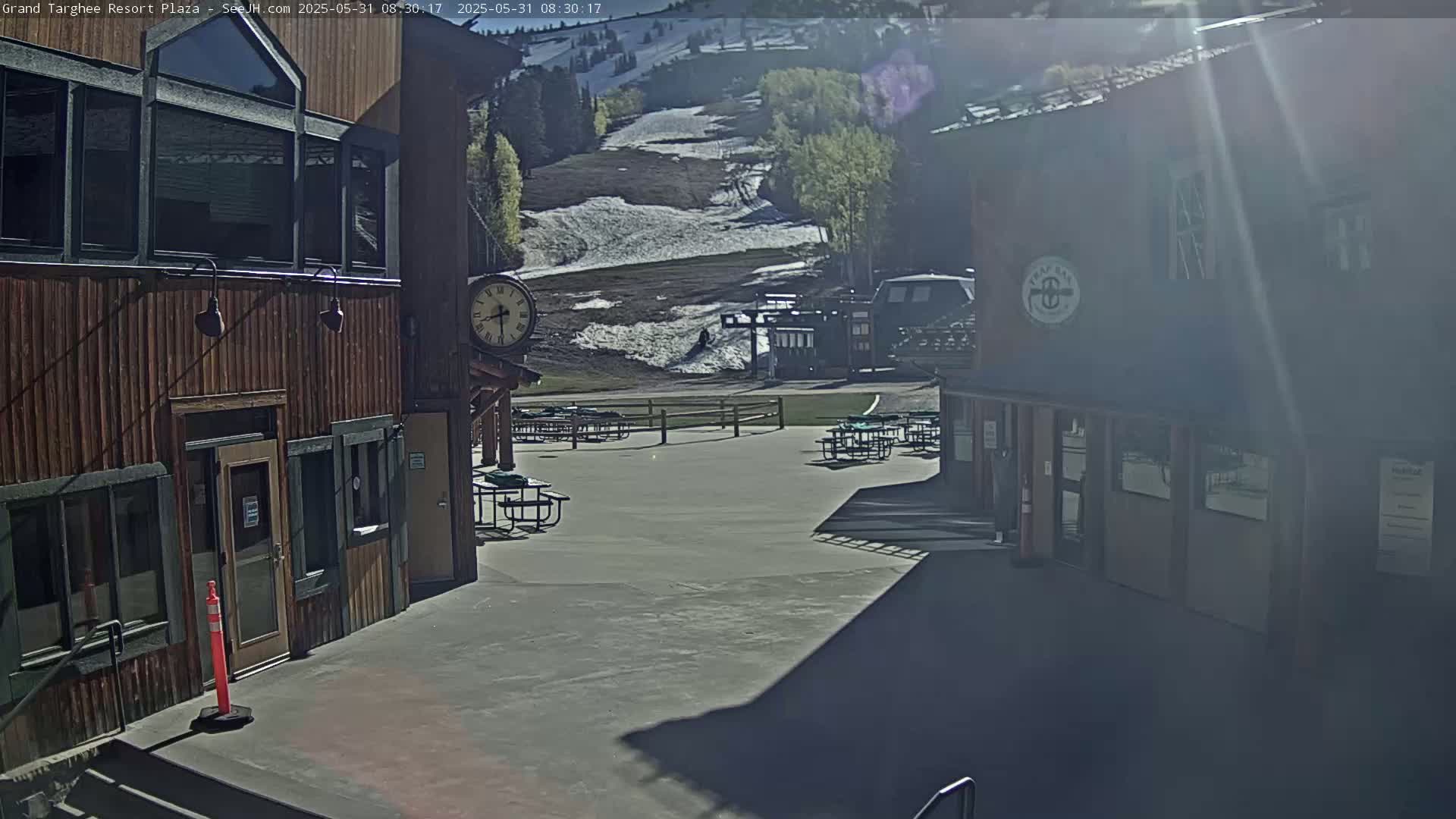 A sunny outdoor plaza with wooden buildings, picnic tables, and a view of a snow-covered ski slope in the background.