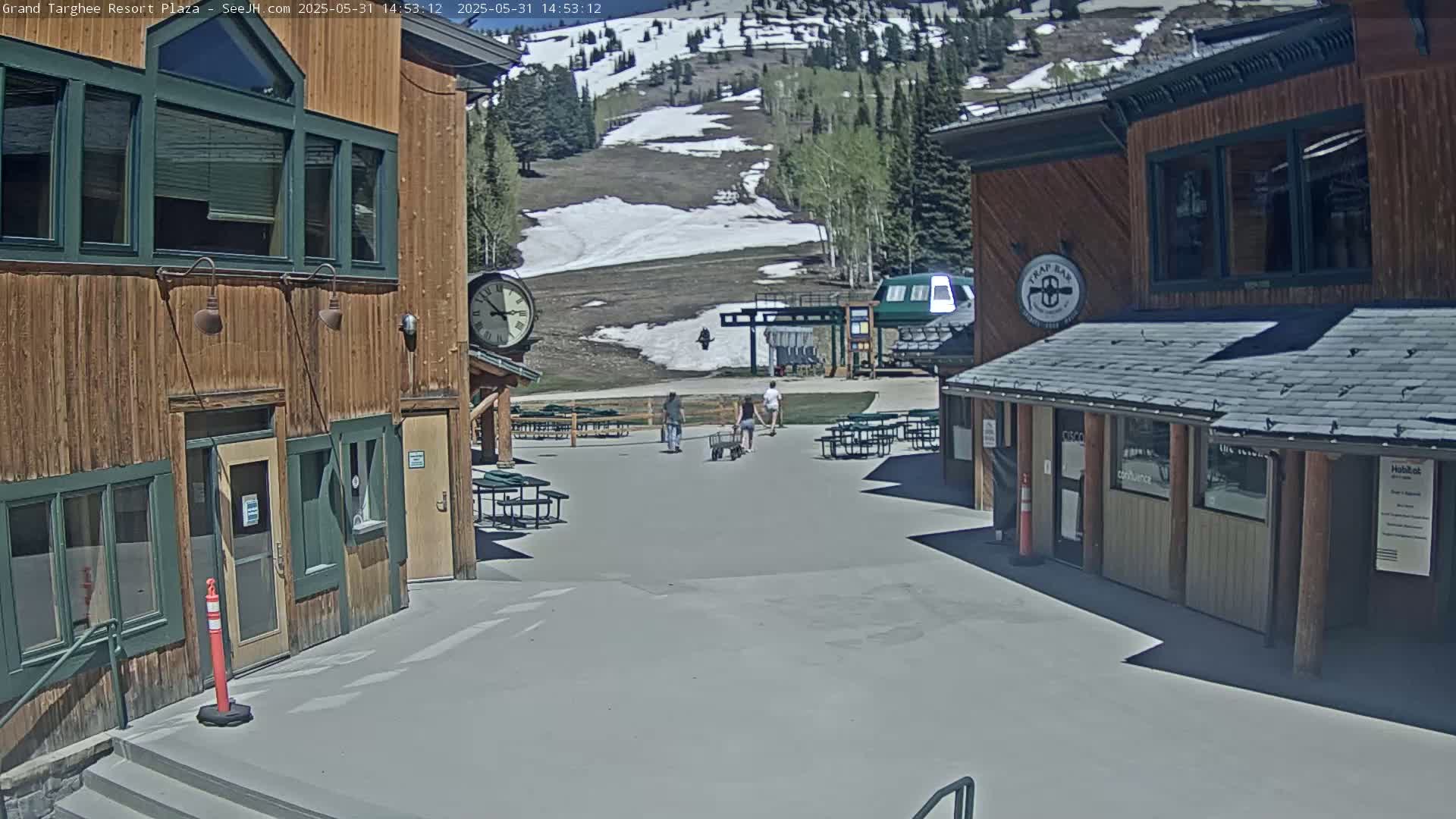 A plaza with two wooden buildings, a ski lift in the background, and three people walking, on a sunny day with some remaining snow on the mountain.