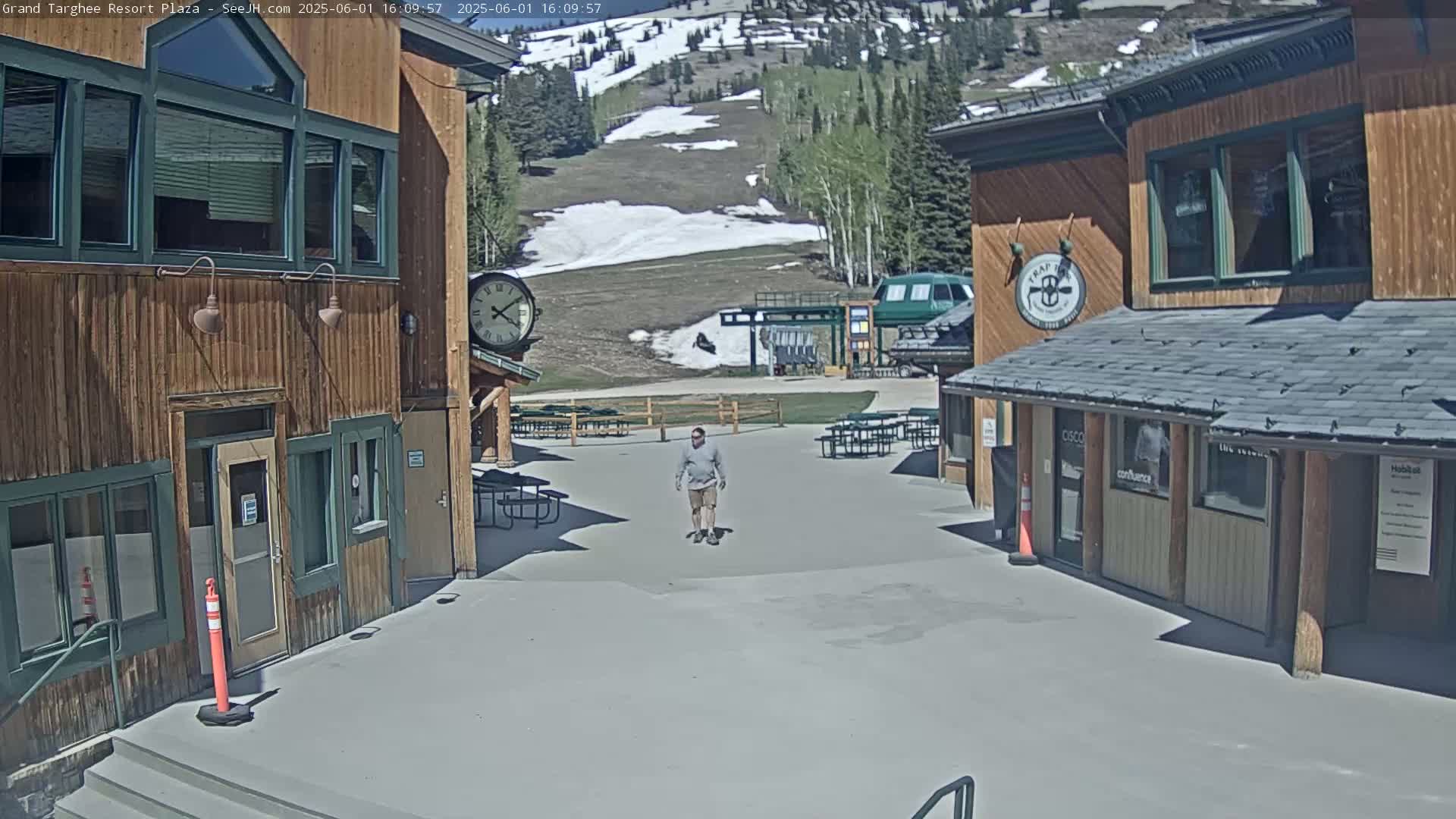 A man walks across a plaza between two wood-sided buildings on a sunny day with patches of snow visible on a nearby mountainside.