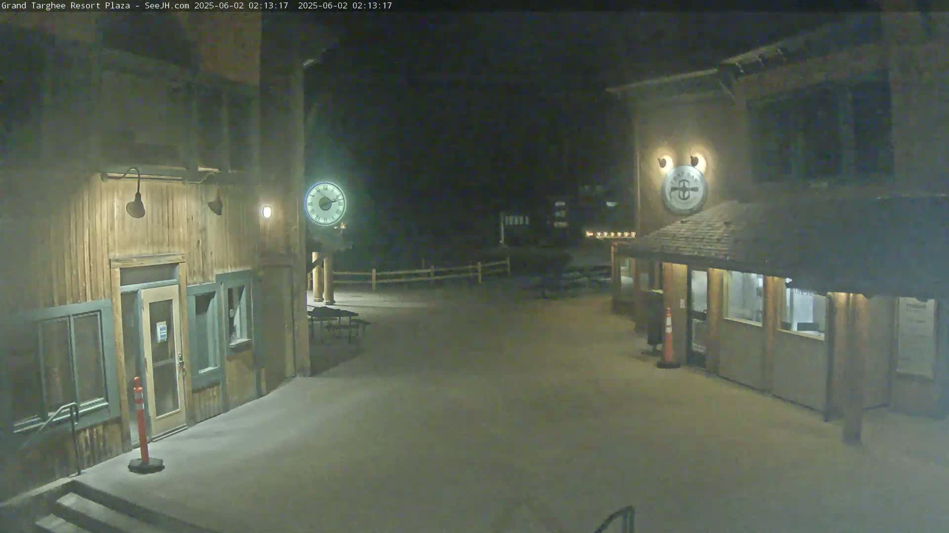 A nighttime view of a plaza with two wooden buildings, outdoor seating, and a fence.