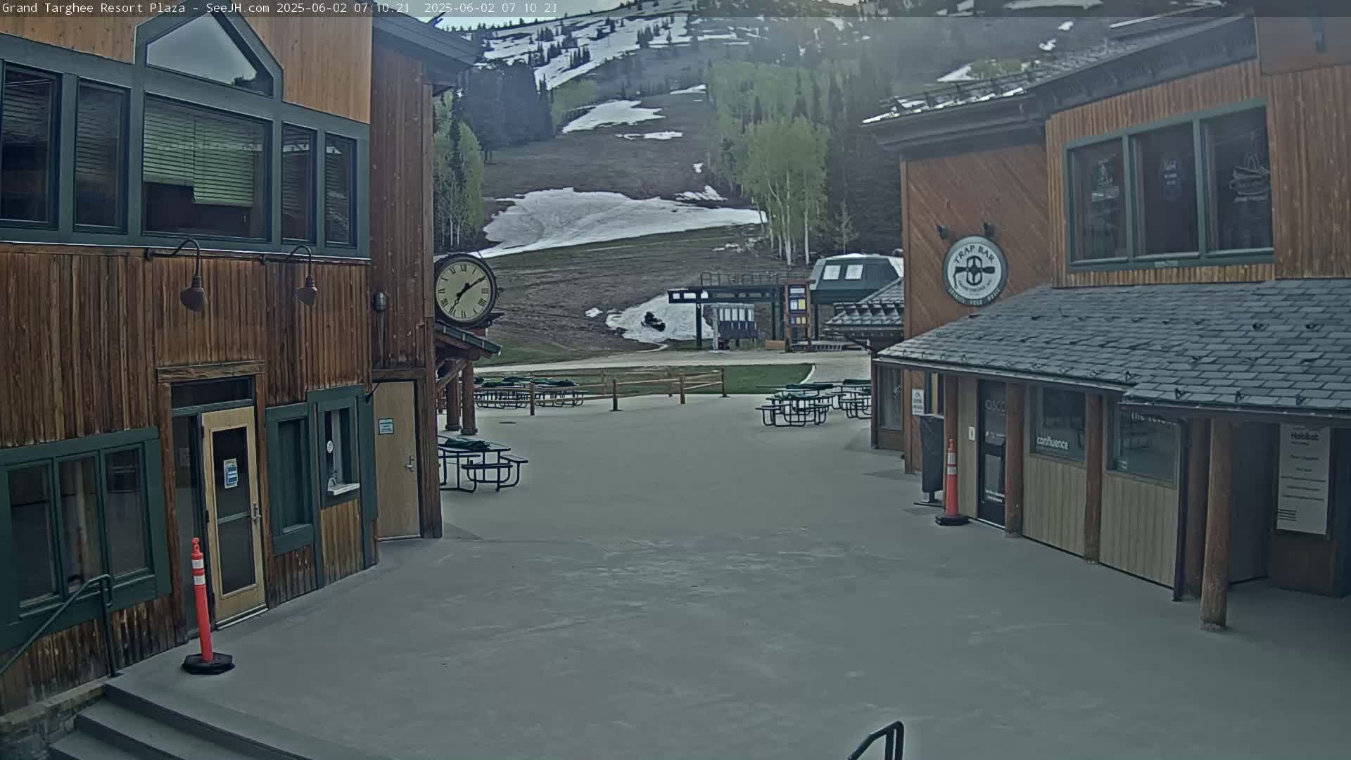 A plaza with wooden buildings and outdoor seating overlooks a ski slope with patches of remaining snow under a mostly clear sky.