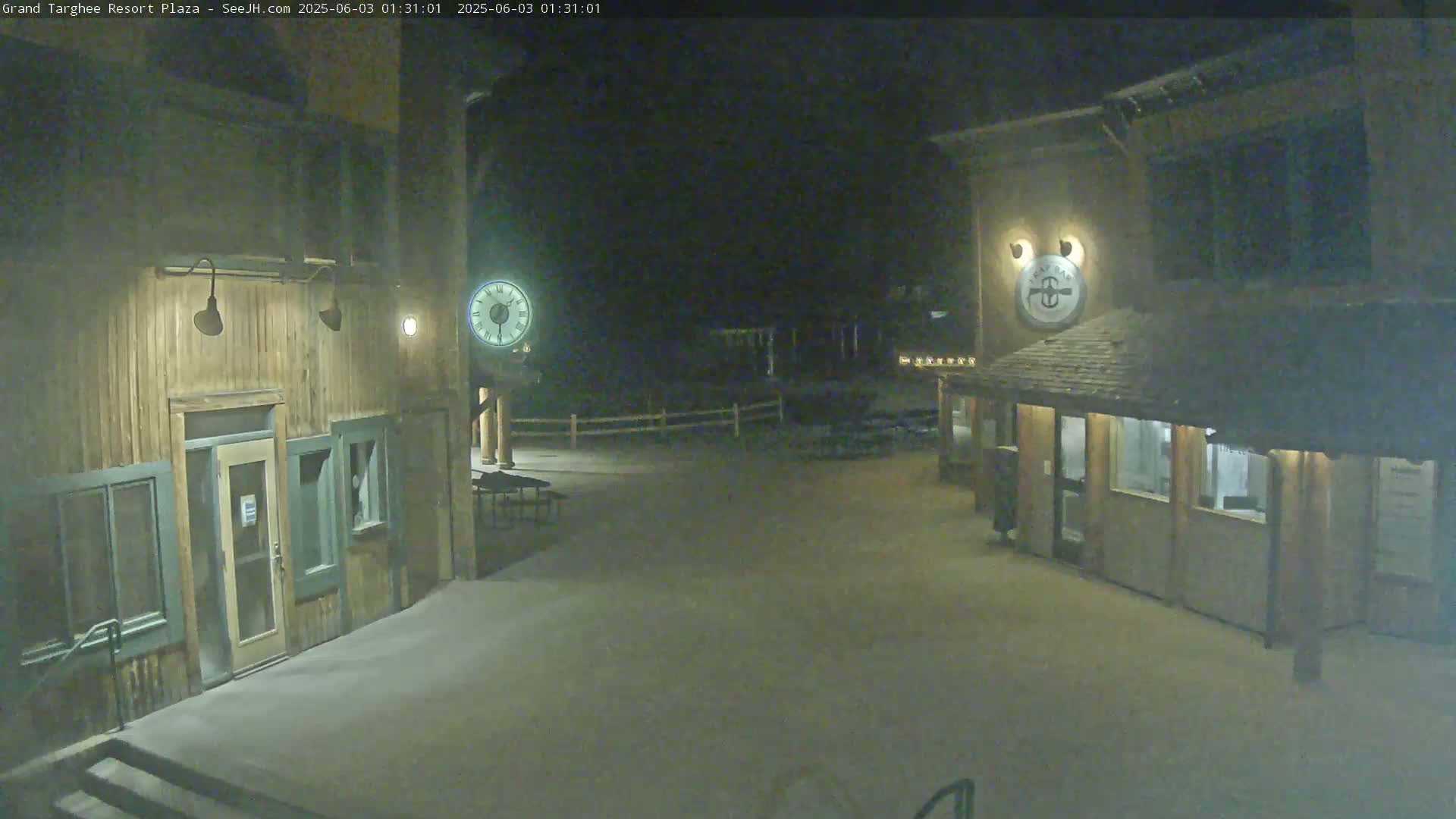 A nighttime view of a snow-covered plaza with two wooden buildings, one featuring a large clock.