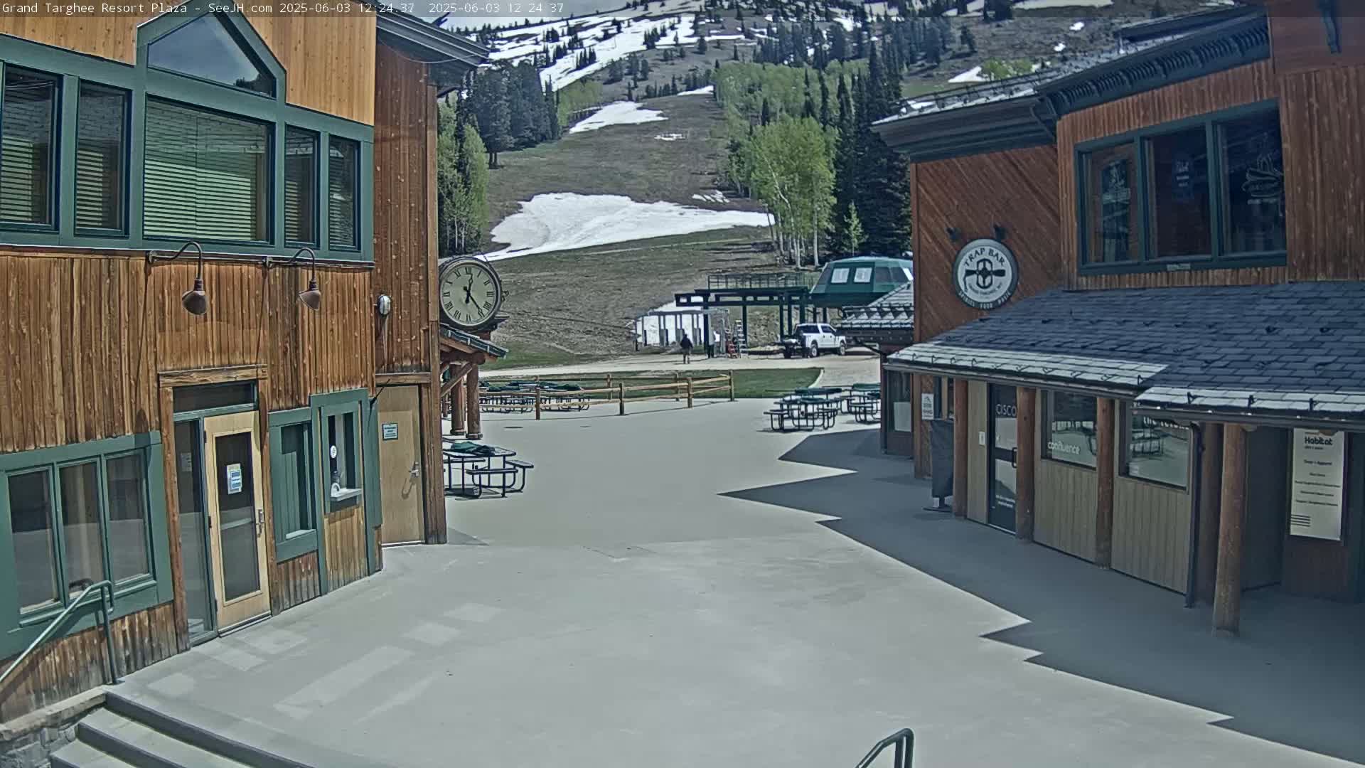 A plaza with wooden buildings and outdoor seating overlooks a ski slope with patches of remaining snow under a sunny sky.