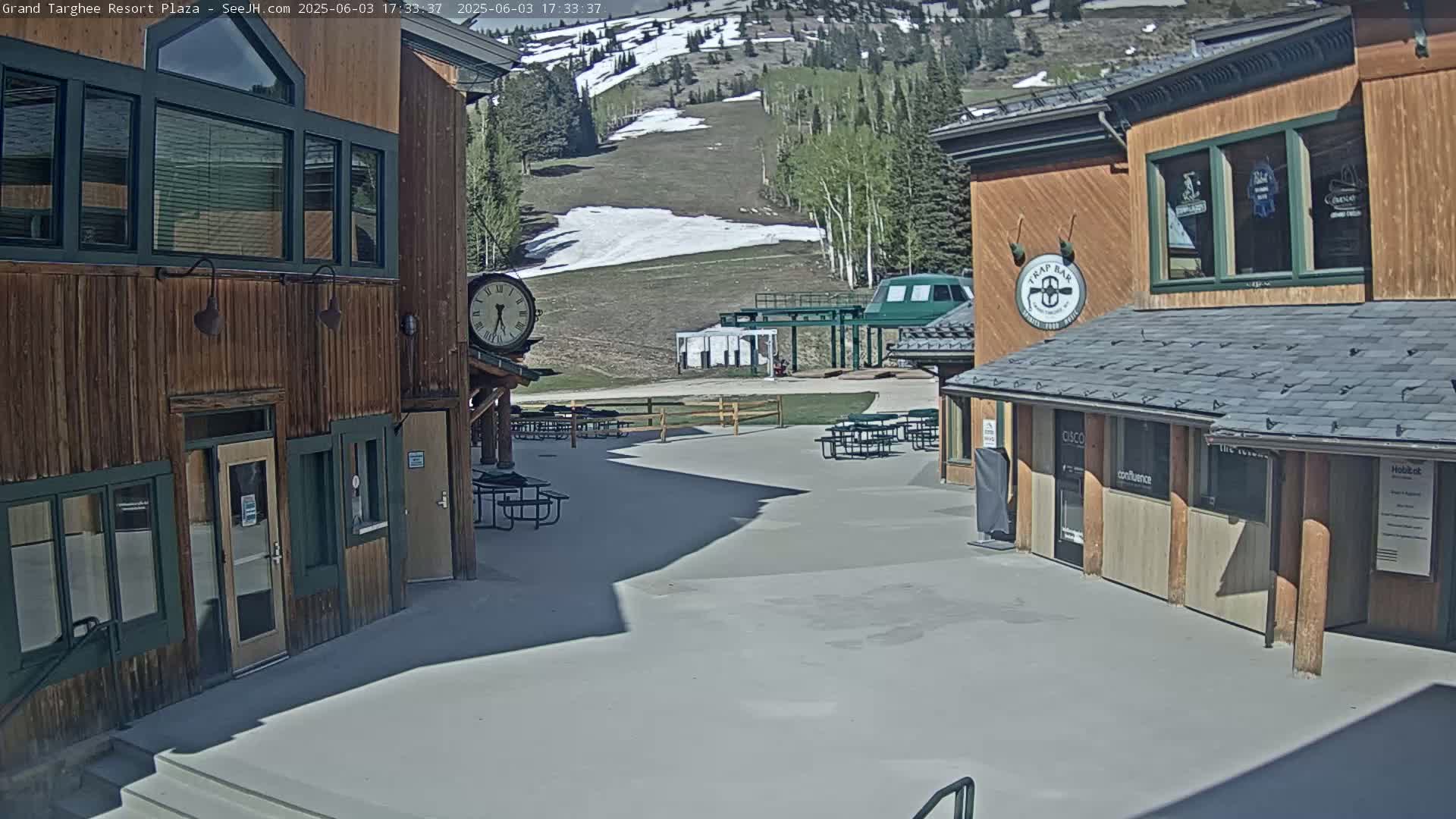 A plaza with wooden buildings, picnic tables, and a ski lift in the background on a sunny day with some remaining snow patches on the mountain.