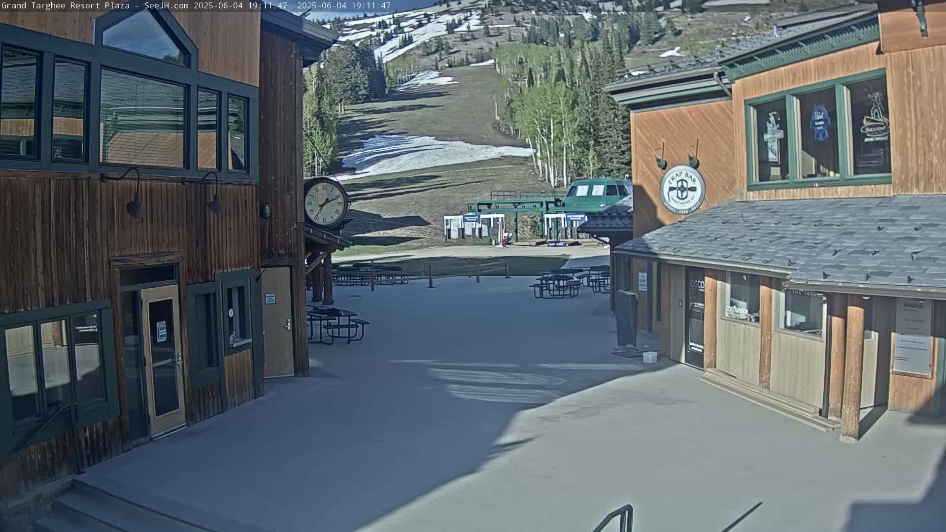 A sunny outdoor plaza with two wooden buildings, picnic tables, and a ski lift in the background.