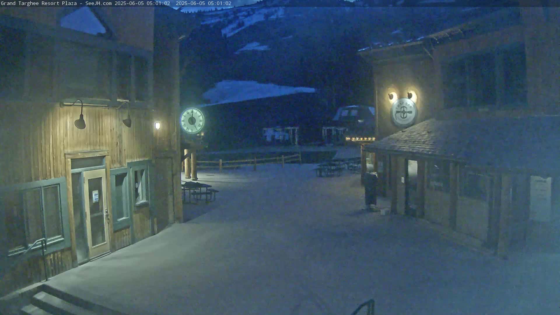 A snow-covered plaza at night is illuminated, showing two wooden buildings with outdoor seating and a large clock.