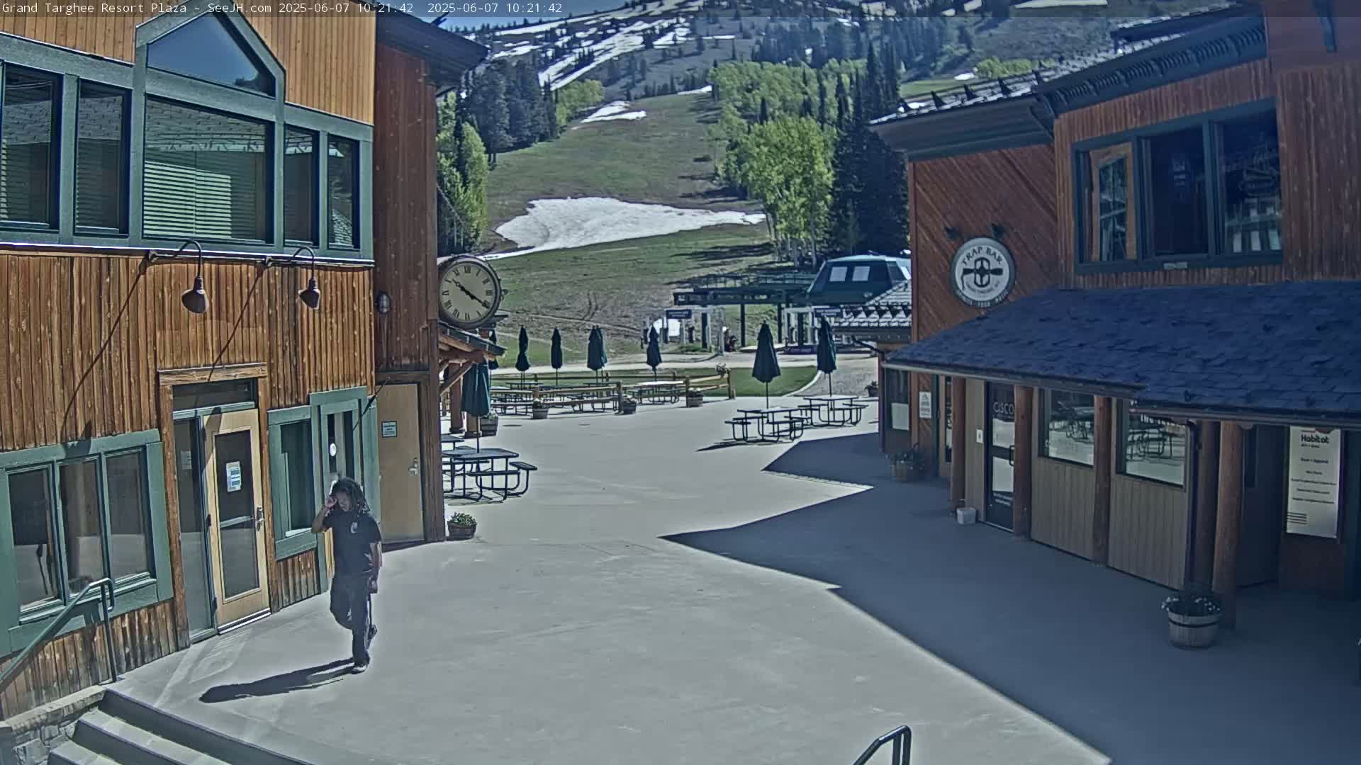 A sunny plaza with wooden buildings, outdoor seating, and a person walking, overlooks a grassy hillside with patches of snow.