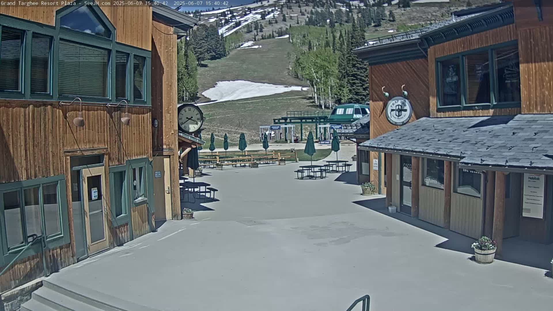 A plaza with several outdoor tables under green umbrellas sits between two wooden buildings on a sunny day, with a ski slope and a small patch of snow visible in the background.