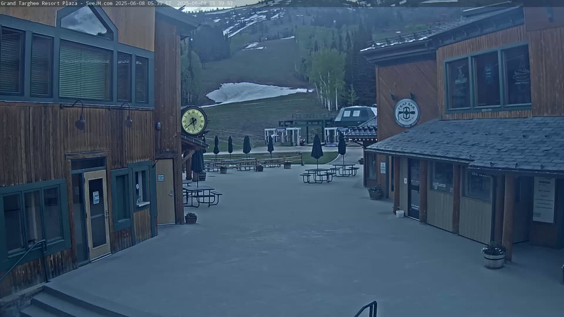 A plaza with several wooden buildings and picnic tables under umbrellas, overlooking a grassy hillside with some remaining snow, under a partly cloudy sky.