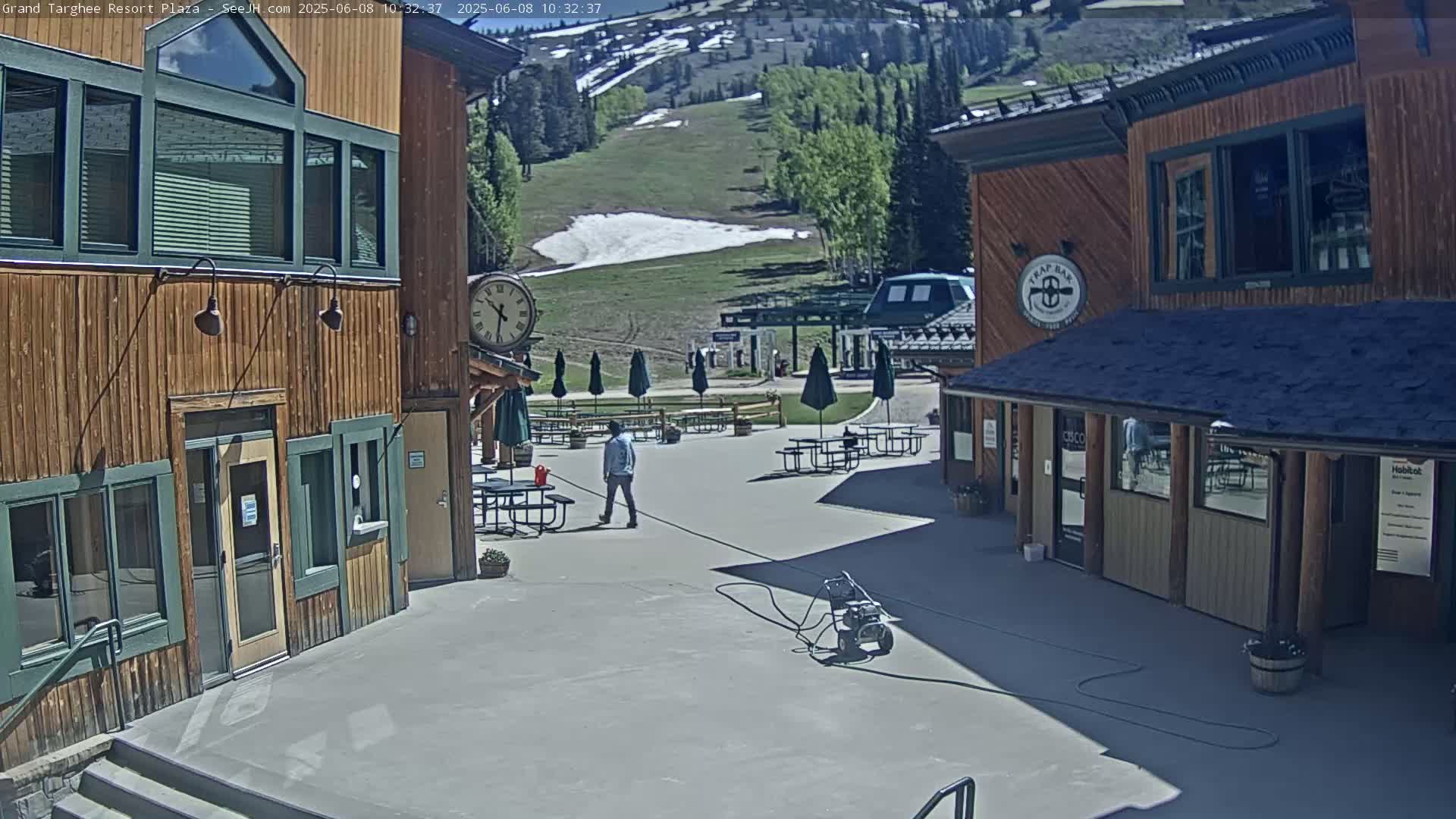 A sunny outdoor plaza with wooden buildings, a person walking, several picnic tables with umbrellas, and a grassy hillside with some remaining snow in the background.
