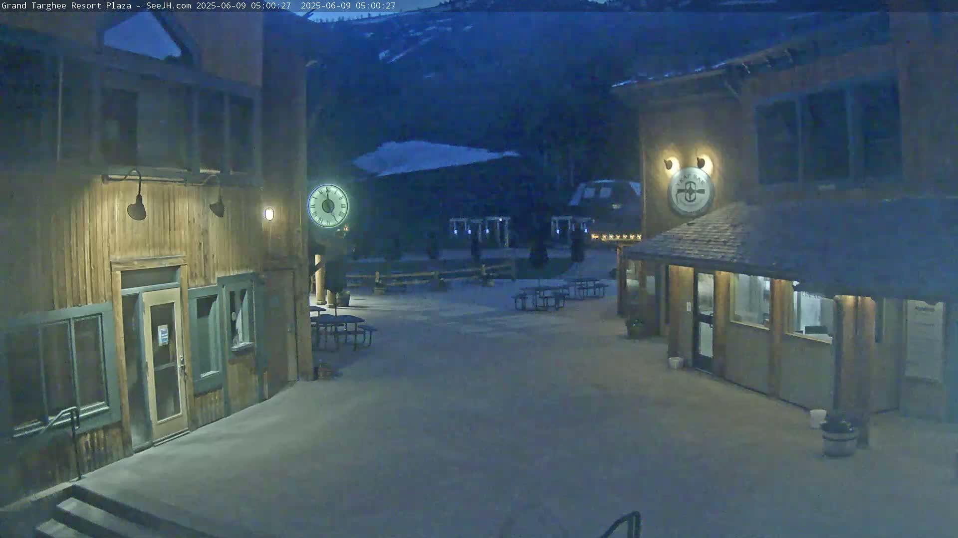 A snow-covered plaza at night, between two wooden buildings with outdoor seating, under a dark sky.
