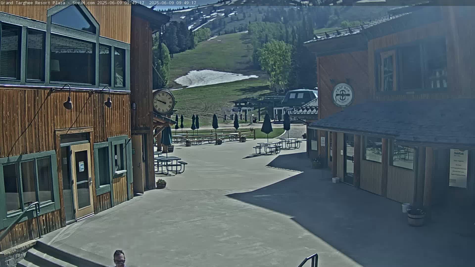 A plaza with wooden buildings and picnic tables under umbrellas, overlooking a grassy hillside with a patch of remaining snow, on a sunny day.