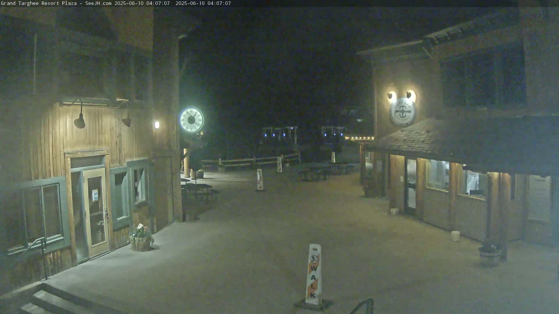 A nighttime, outdoor view of a plaza with two wooden buildings, several empty picnic tables, and a "walk" sign.