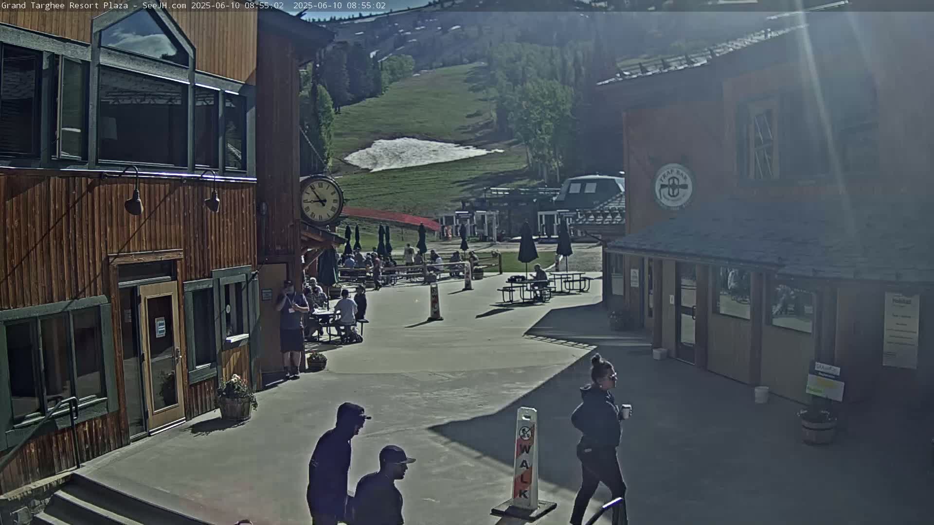 A sunny outdoor plaza scene shows several people sitting at tables near wooden buildings, with a grassy hill and a small patch of snow visible in the background.