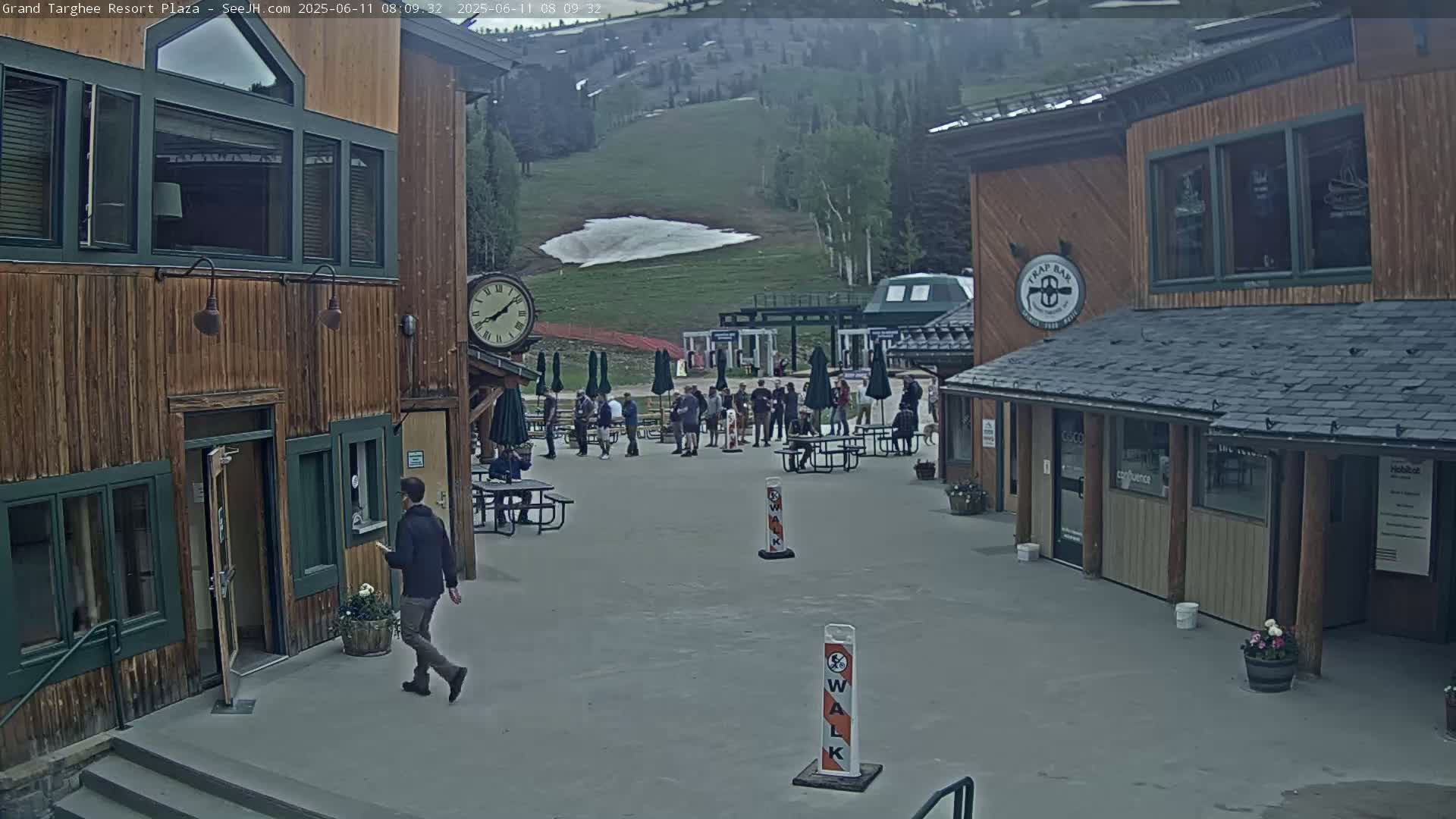 A plaza with several wooden buildings, outdoor seating, and a scattering of people, overlooks a grassy hillside with a small patch of snow, under overcast skies.