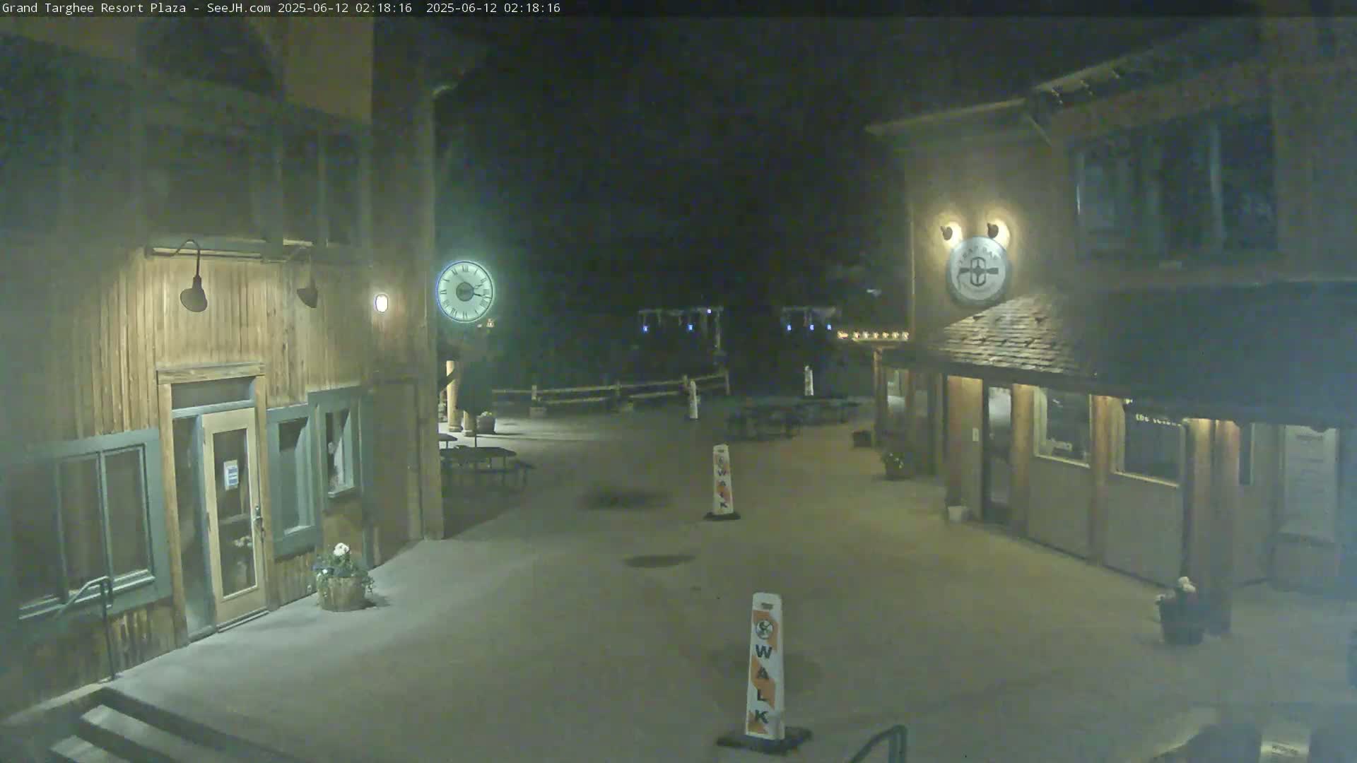 An empty plaza at night, illuminated by artificial lights, features two wooden buildings and several outdoor tables.