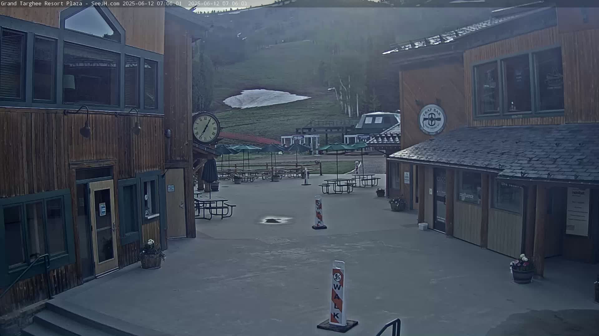 A plaza with several wooden buildings, picnic tables, and umbrellas sits under a hazy sky, with a patch of snow visible on a hillside in the background.