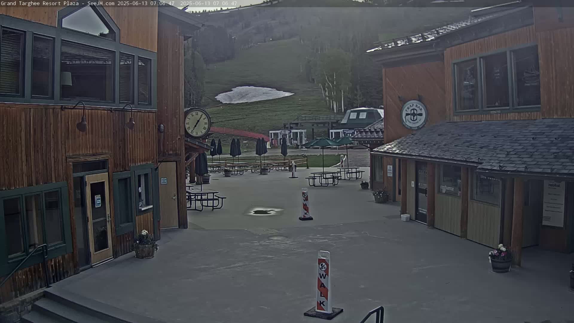 A plaza with wooden buildings, picnic tables, and umbrellas, overlooks a grassy hillside with a small patch of snow, under an overcast sky.