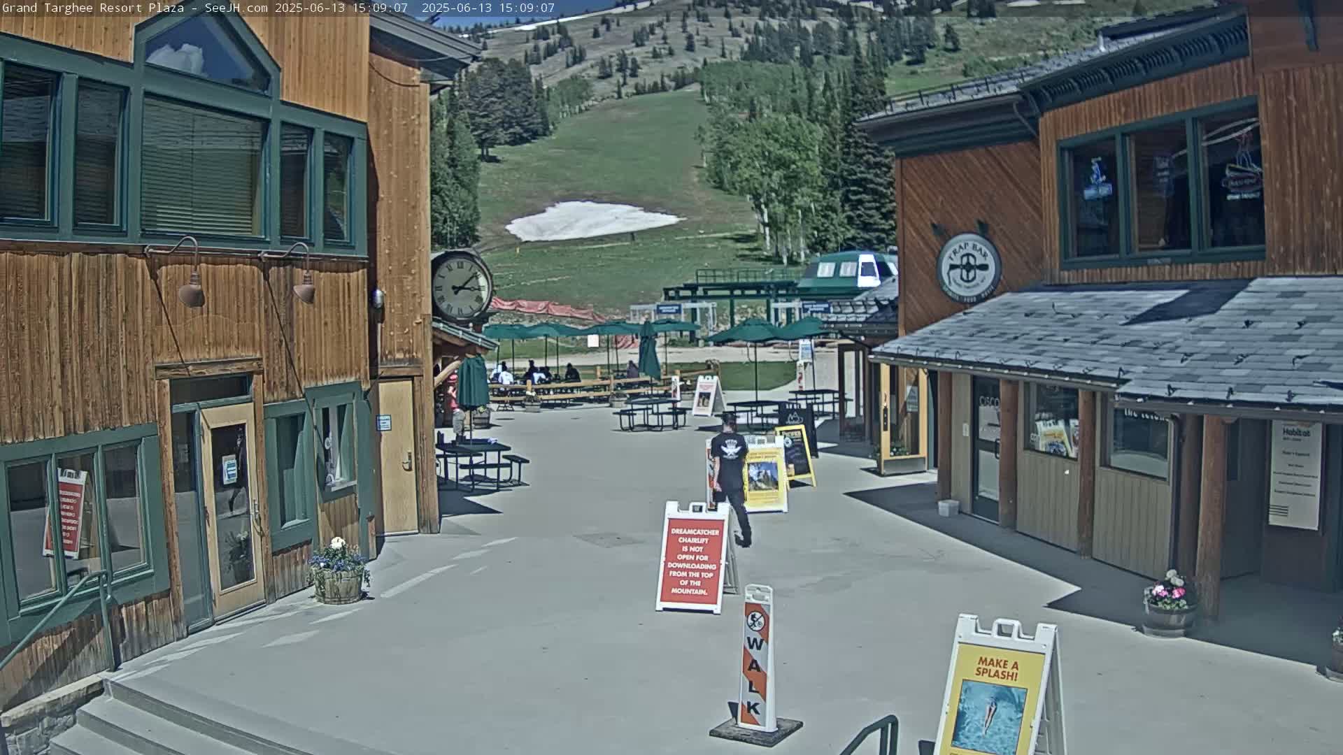 A plaza with wooden buildings, outdoor seating areas, and a partially snow-covered hillside in the background on a sunny day.