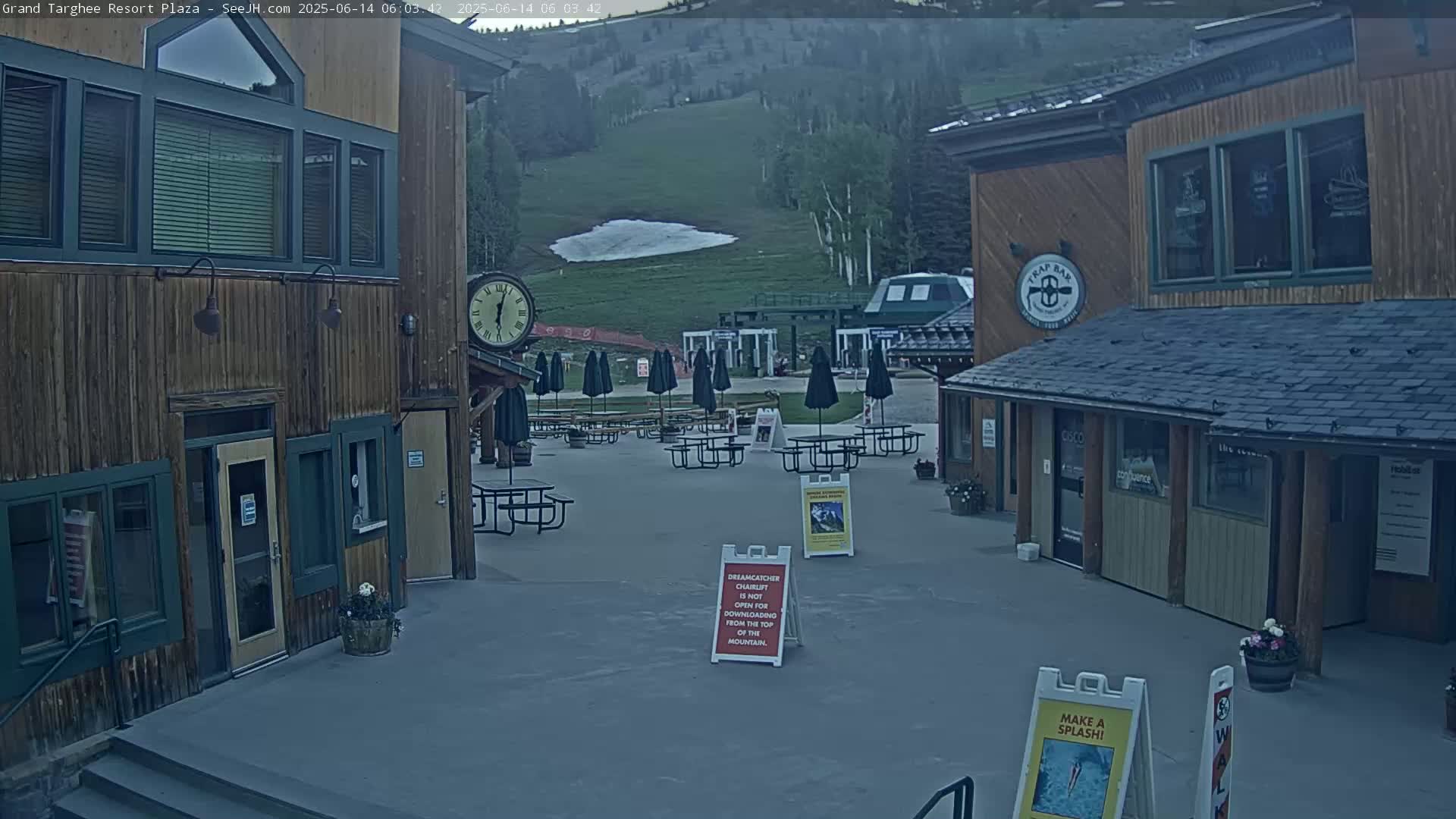 An outdoor plaza with wooden buildings, picnic tables under umbrellas, and a mountain in the background under overcast skies.