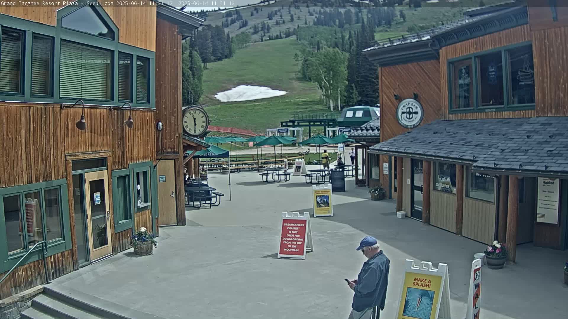 A plaza with wooden buildings, outdoor seating, and umbrellas overlooks a grassy hillside with a small patch of snow, under a sunny sky.