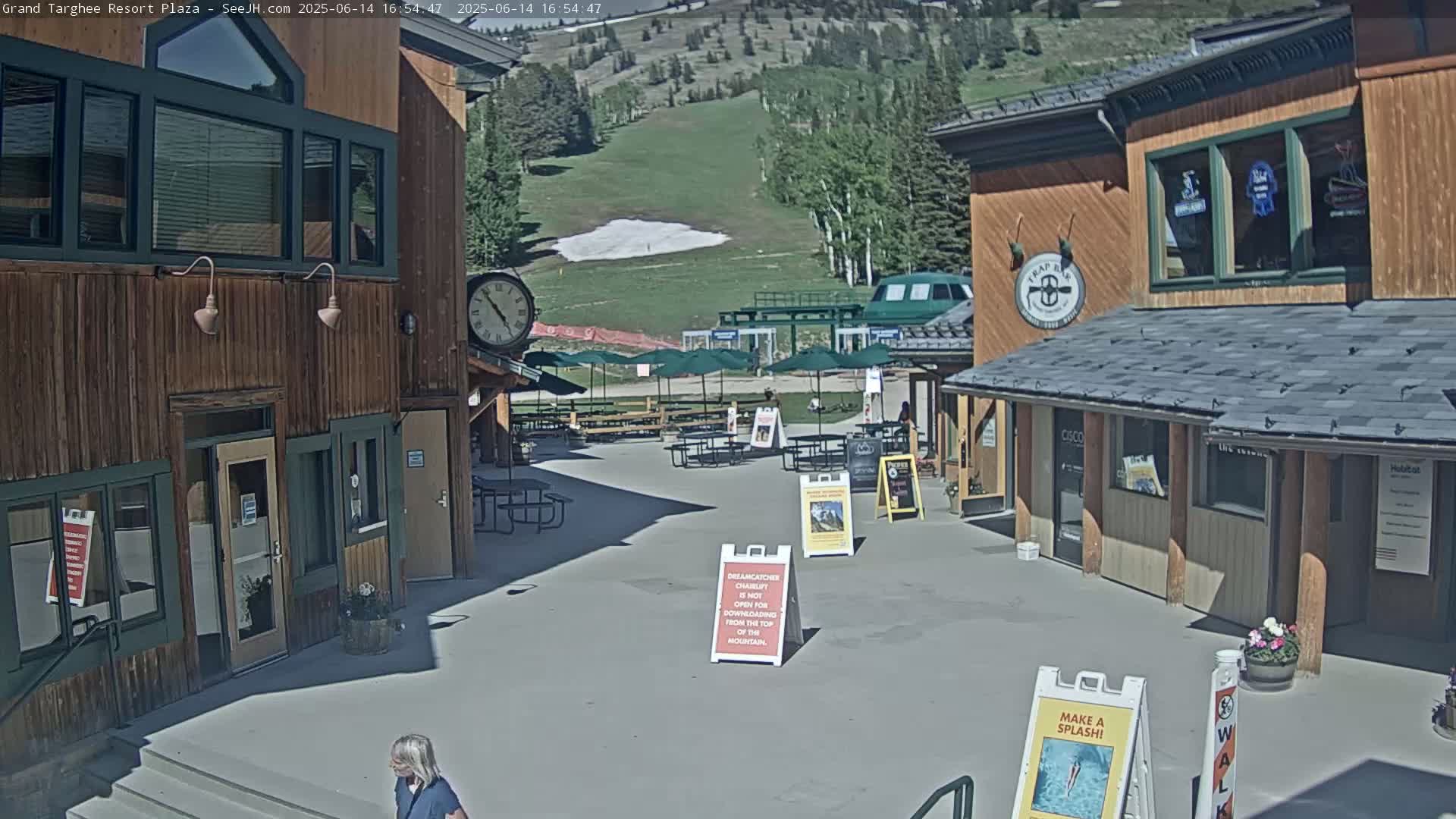 A plaza with wooden buildings, outdoor seating, and umbrellas overlooks a grassy hillside with a small patch of snow, under a sunny sky.