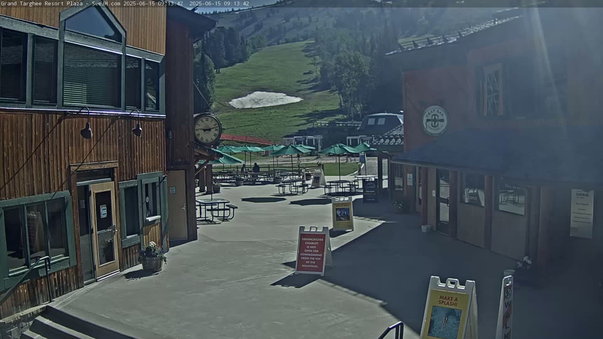 A sunny outdoor plaza with wooden buildings, patio furniture under umbrellas, and a grassy hill in the background.