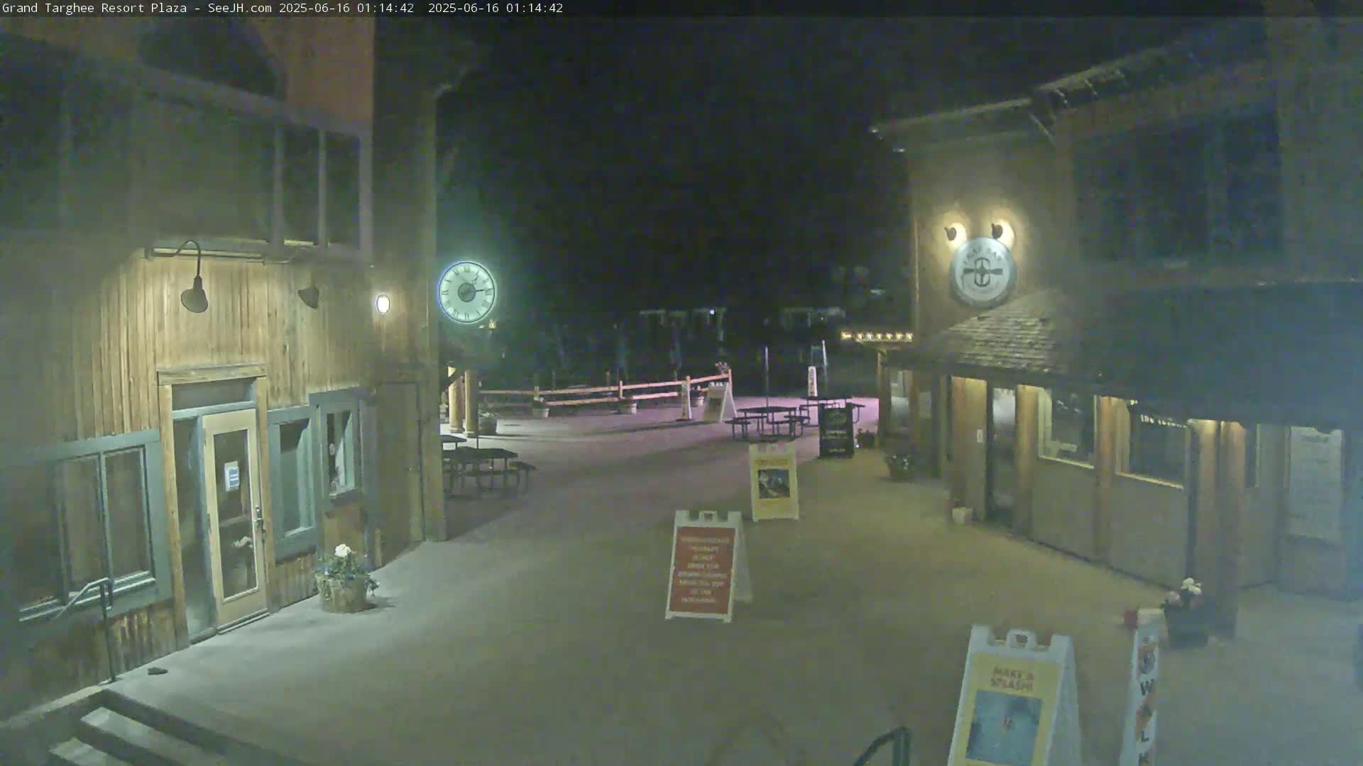 A nighttime view of a plaza with two wooden buildings, outdoor seating, and a clock.