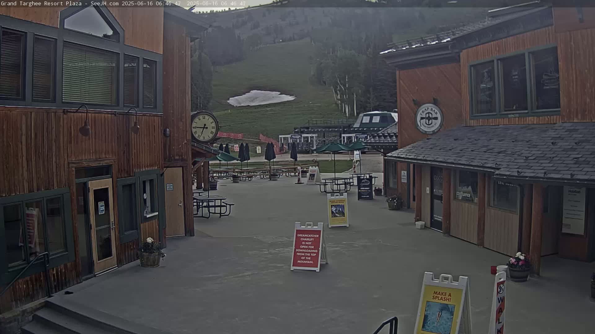 An outdoor plaza with wooden buildings, picnic tables under umbrellas, and a grassy hill with a small patch of snow in the background under an overcast sky.