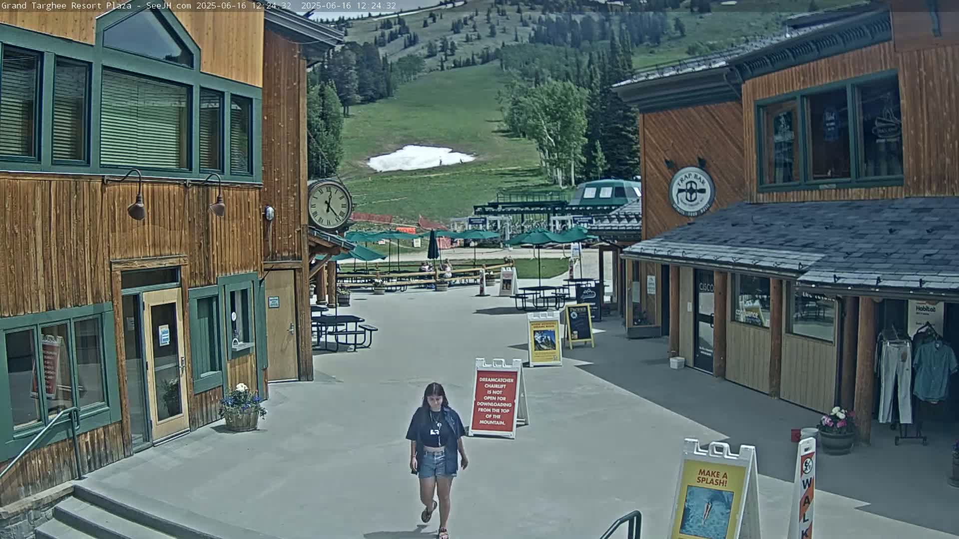A plaza with wooden buildings, outdoor seating, and a person walking, under a sunny sky with a small patch of snow visible on a hillside in the background.
