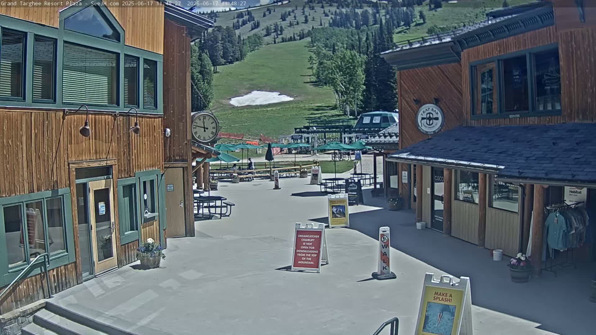A sunny plaza with wooden buildings, outdoor seating, and a grassy hillside in the background.