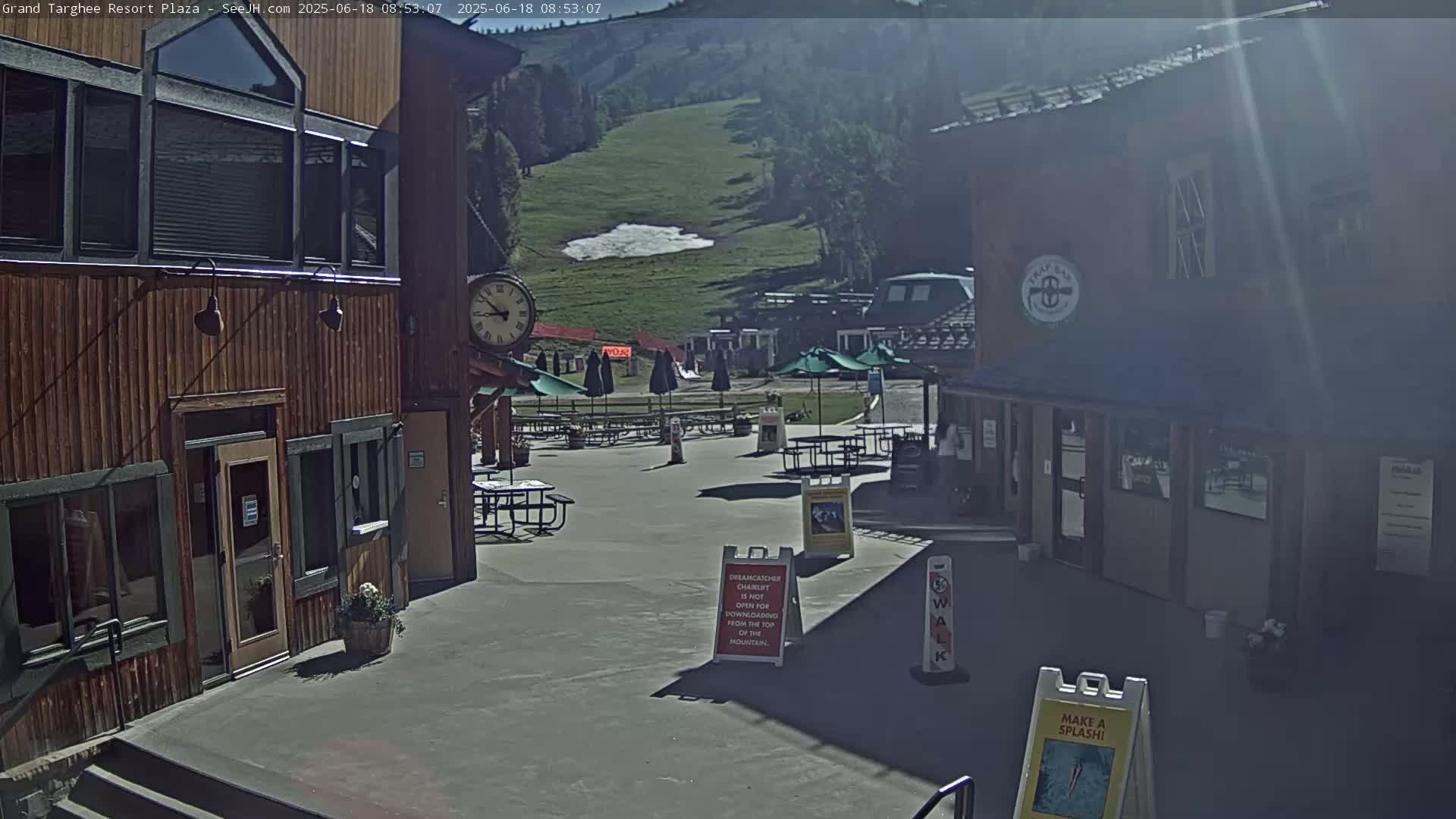 A sunny plaza with wooden buildings, outdoor seating, and a grassy hillside with a small patch of snow in the background.