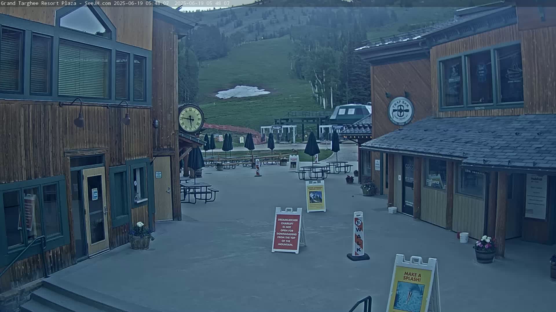 An outdoor plaza with wooden buildings, picnic tables under umbrellas, and a grassy hillside in the background under a twilight sky.