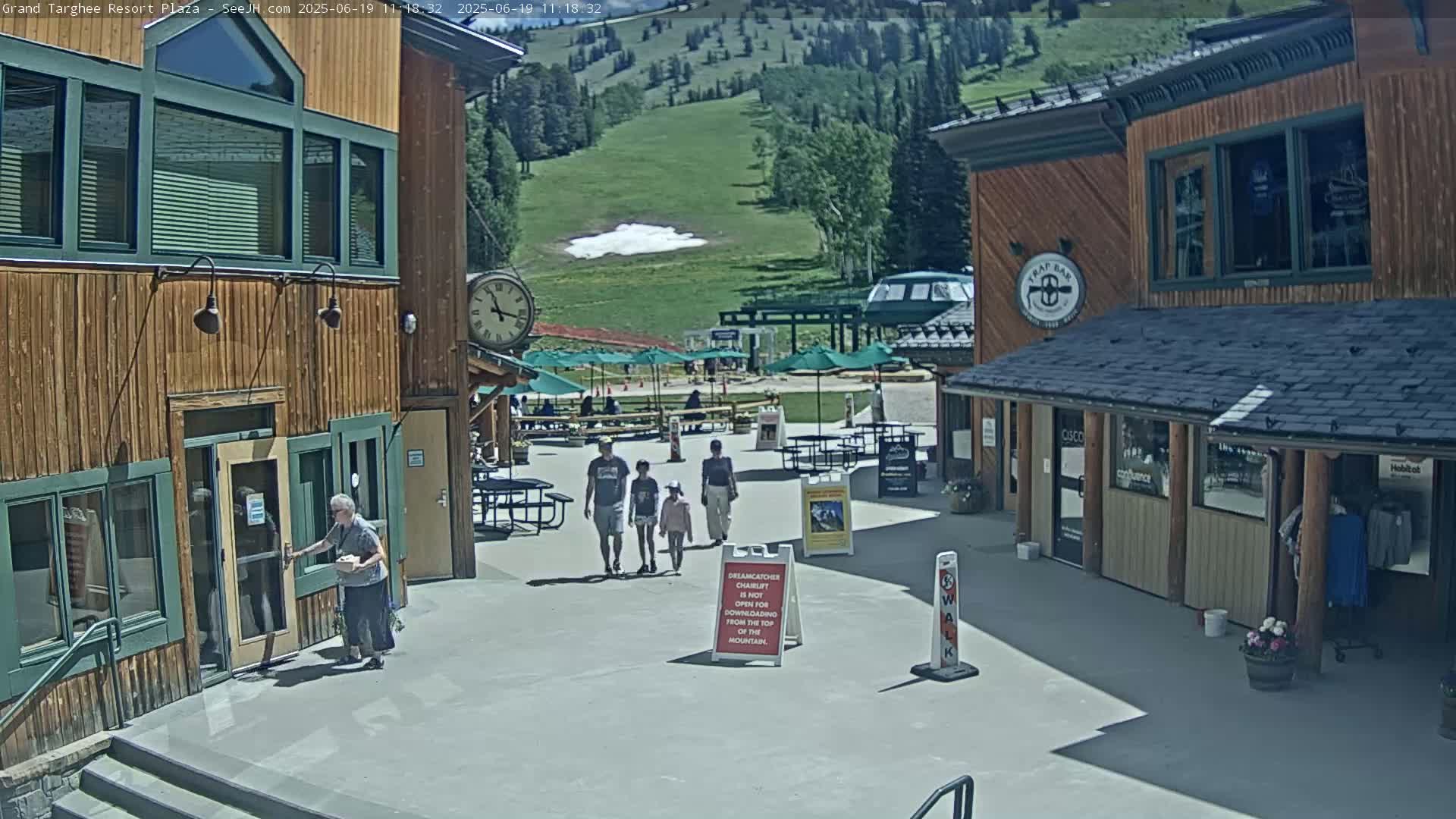A sunny plaza with wooden buildings, a family walking, and a grassy mountainside in the background.