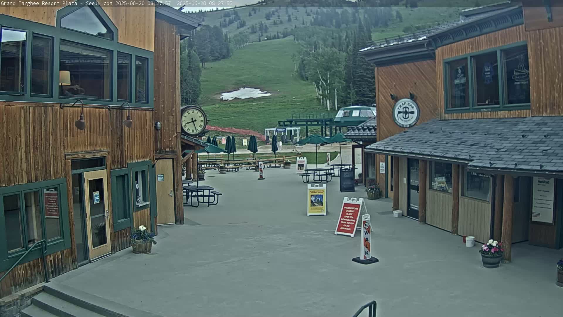 An outdoor plaza with wooden buildings, picnic tables under umbrellas, and a grassy hillside in the background under an overcast sky.