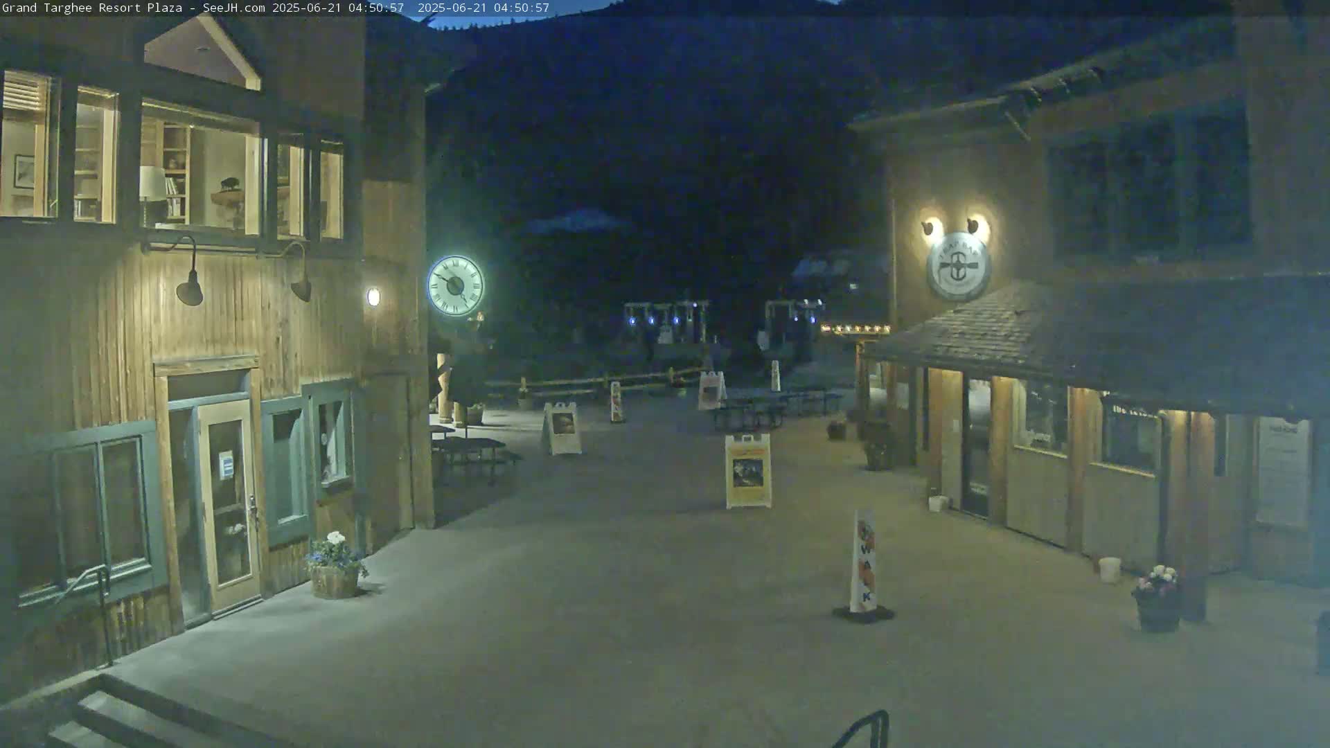 A nighttime view of a plaza with two wooden buildings, outdoor seating, and a large clock, all situated against a dark mountain backdrop.