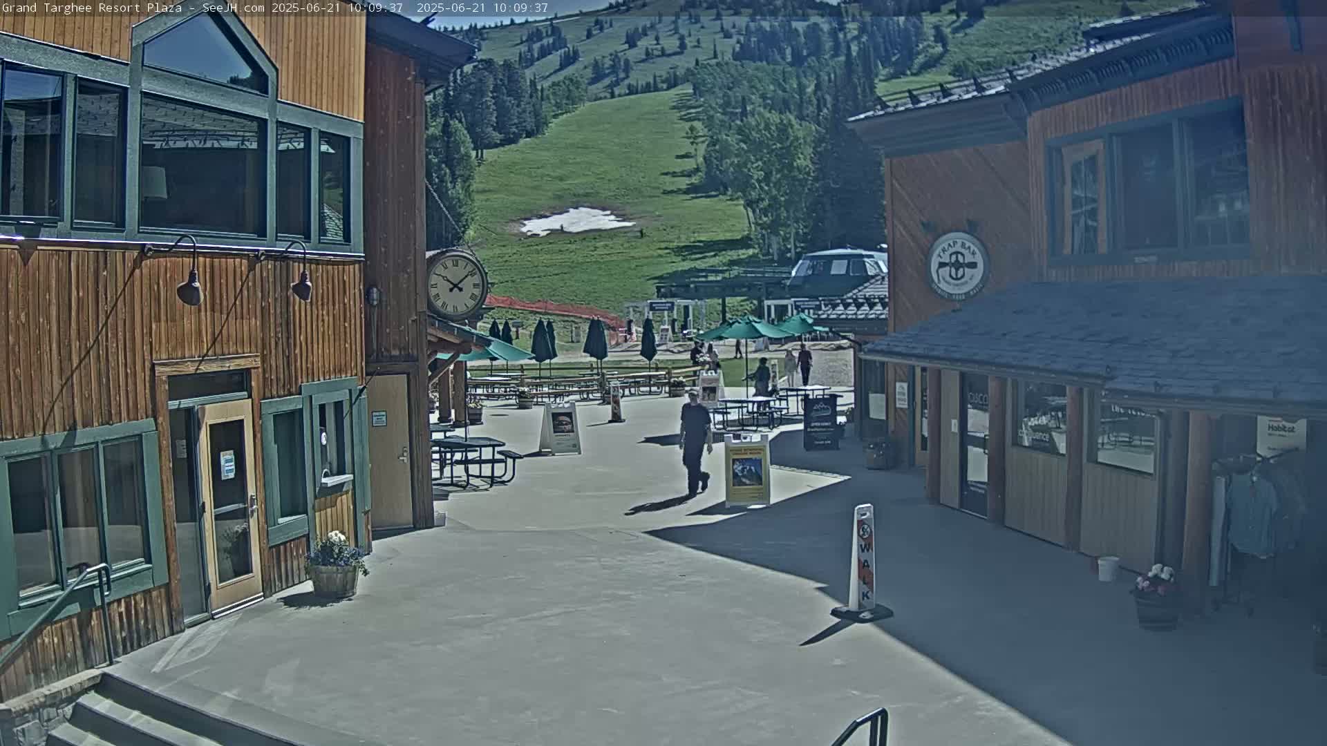 A sunny plaza with wooden buildings, outdoor seating, and a grassy hillside in the background.