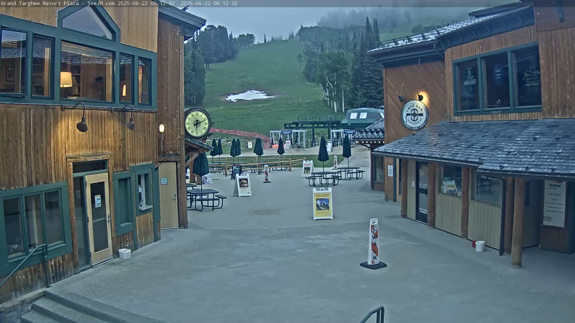 An overcast day reveals a plaza with several wooden buildings, picnic tables, and umbrellas, overlooking a grassy hillside with a ski lift.